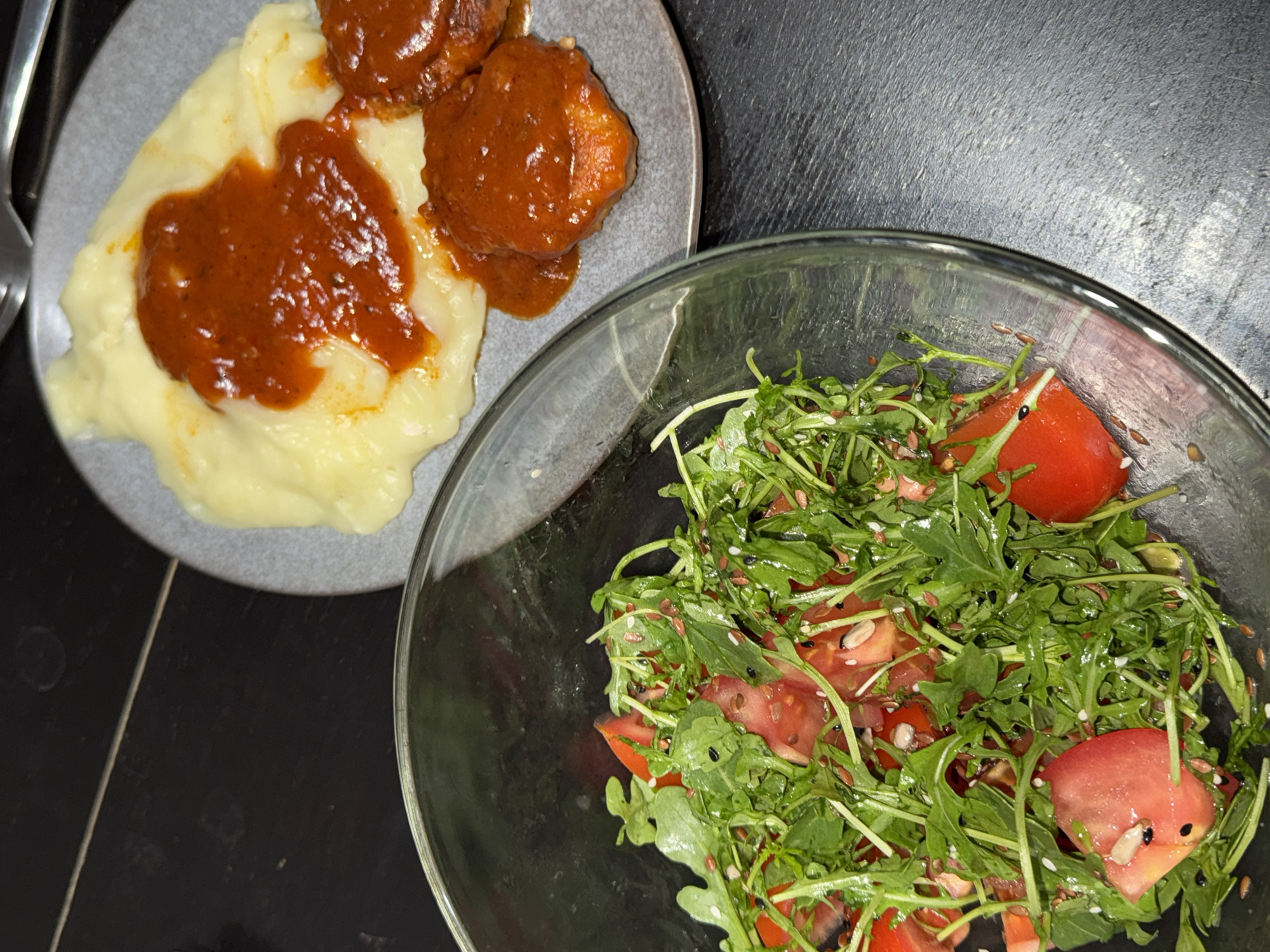 The image shows a meal consisting of mashed potatoes topped with a dark red sauce and meatballs on a gray plate, accompanied by a salad in a clear glass bowl. The salad appears to contain tomatoes and leafy greens, possibly arugula, sprinkled with seeds. The food is placed on a dark, possibly metallic, surface. The location, Perm, Russia, is not discernible from the image.