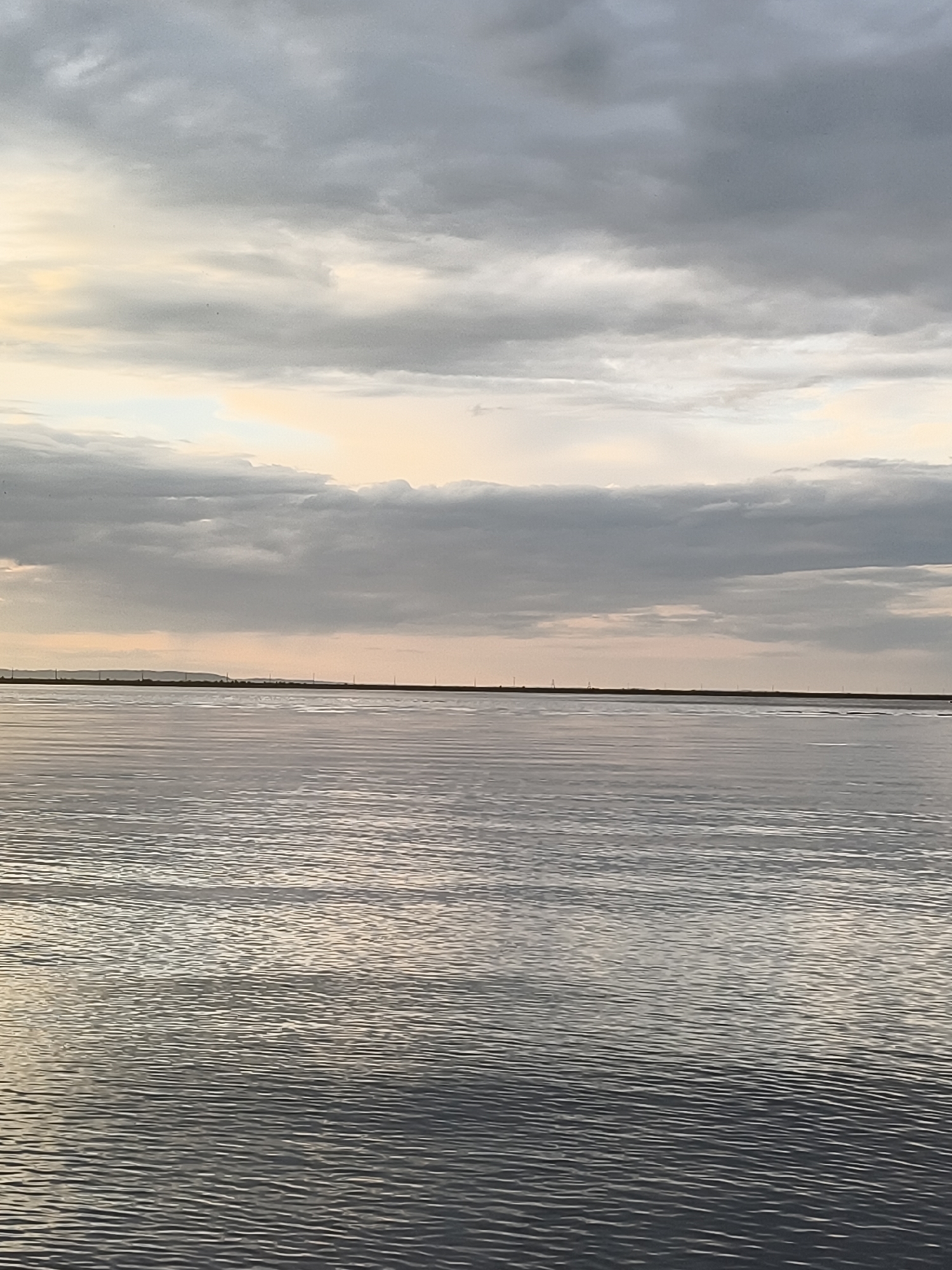 This media file depicts a wide, calm body of water under an overcast sky. The water's surface is rippled, reflecting the subdued light. In the distance, a low, dark horizon line suggests land, with faint, evenly spaced vertical structures that could be power lines or poles. The sky is filled with layered grey clouds, with lighter patches of pale yellow and white indicating the presence of the sun behind the clouds, suggesting either dawn or dusk. The overall mood is serene and slightly melancholic, characteristic of a cloudy day. The location is identified as Chaikovsky, Russia. No people, objects, or text are clearly visible in the image.