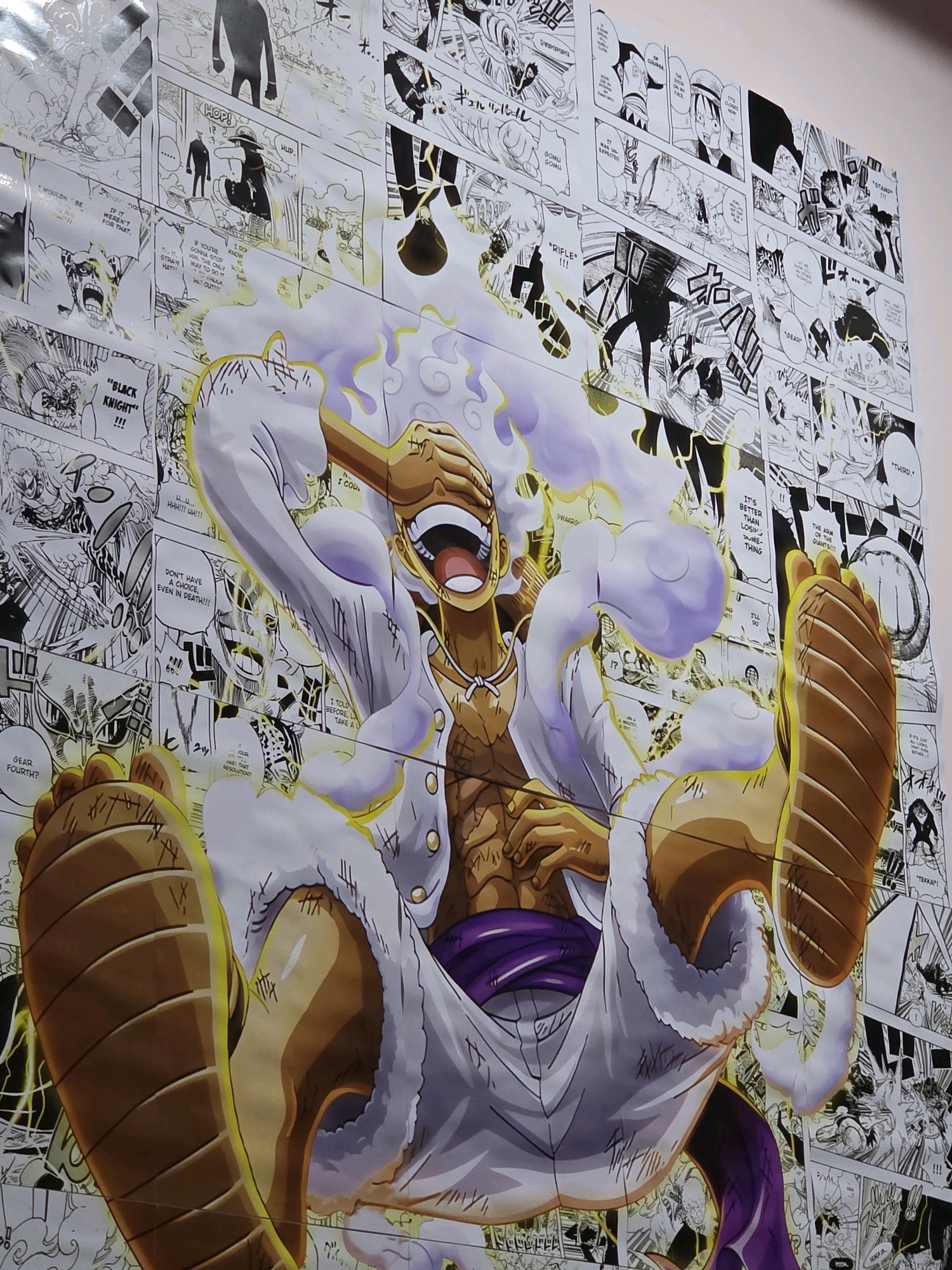 The image is a photograph of a large poster featuring characters from the anime and manga series One Piece. The central figure is Monkey D. Luffy in his Gear Fourth form, characterized by his large, inflated physique, white fur-trimmed attire, and golden aura. He is depicted mid-action, with his mouth wide open in a roar and his eyes covered by his hand. The background of the poster is a collage of black and white manga panels from the series, showcasing various scenes and characters. The poster appears to be mounted on a wall. The location of the poster in Lakkarajupalem, India is not discernible from the image itself.