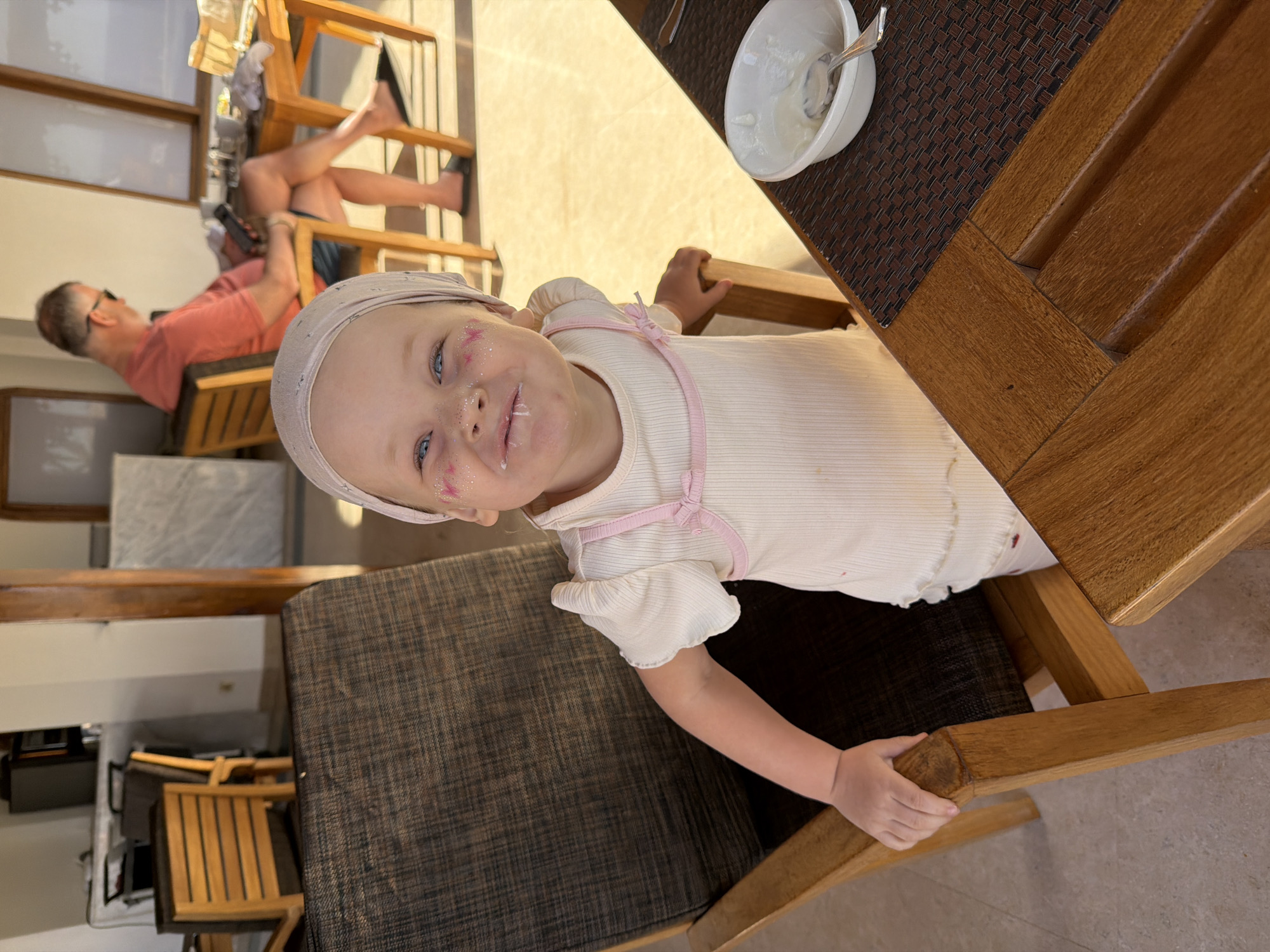 The media file, rotated 90 degrees clockwise from its intended orientation, captures a lively scene in a brightly lit indoor or semi-outdoor dining/lounge area, likely in Sharm El Sheikh, Egypt.
The primary subject is a young child, appearing to be a toddler, positioned in the foreground and looking directly at the camera with a wide, joyful smile. The child has striking blue eyes, is wearing a light-colored, possibly white or cream, short-sleeved top with pink strap details, and a light-colored headscarf. Their face is playfully messy with a white substance, likely yogurt, around their mouth and chin, and sparkles of glitter adorn their cheeks. The child is seated at a wooden table, holding onto the wooden frame of a chair.
On the table next to the child is a white bowl containing a white, creamy substance, likely more yogurt, with a spoon resting inside it. A dark, woven placemat is visible under the bowl.
In the background, slightly out of focus, an adult male is seen relaxing. He wears a peach or orange t-shirt and sunglasses, reclining comfortably in a wooden chair with his bare feet propped up on another chair.
The setting suggests a relaxed, casual atmosphere, perhaps a holiday or family meal. The strong, natural sunlight illuminating the scene indicates it is daytime. The overall emotion conveyed by the child's expression is one of happiness and contentment. No visible text is present in the image.