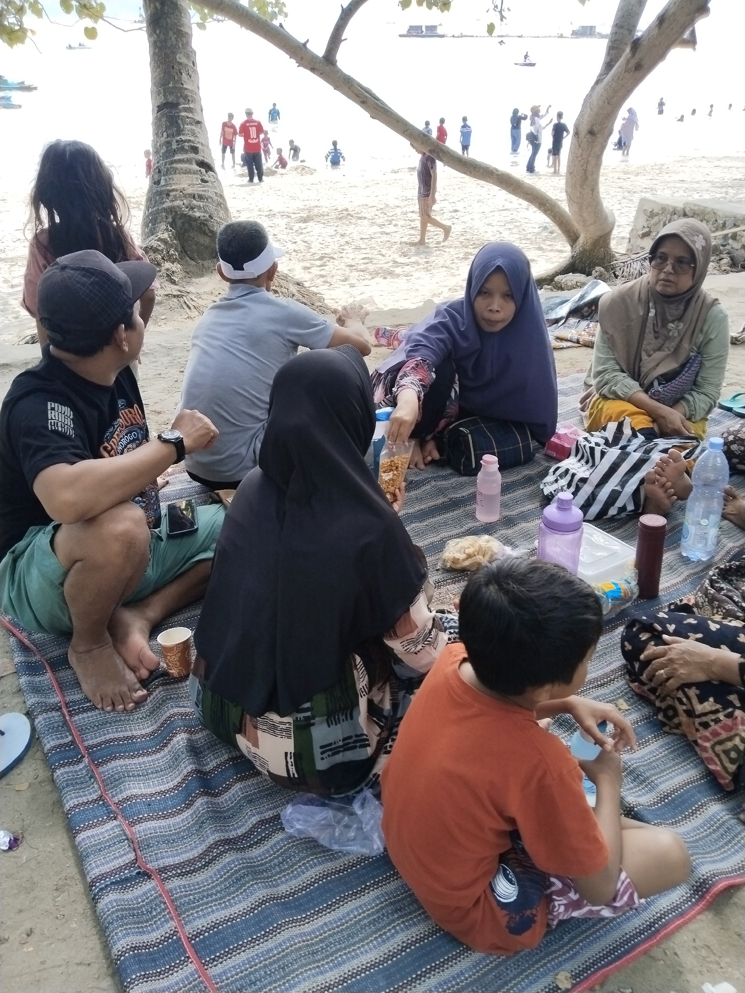 The image captures a scene at the beach in Prigi, Indonesia, likely during daytime. Several individuals are seated on a striped mat on the sand, enjoying a relaxed atmosphere. In the foreground, a man in a black t-shirt and green shorts is seated, with a cup to his left. Next to him, a woman wearing a black hijab is partially visible. A young boy in an orange t-shirt and patterned shorts sits opposite them, holding a bottle. Further back, two women in hijabs are seated, one in a blue hijab and the other in a beige hijab, both appearing to be engaged in conversation or observation. In the background, the beach is populated with other beachgoers, some wading in the water and others walking along the shore. The presence of trees suggests a coastal environment. Various objects are scattered on the mat, including water bottles, food packaging, and a mobile phone, indicating a picnic or gathering. The overall impression is one of a casual outing at a popular beach destination.