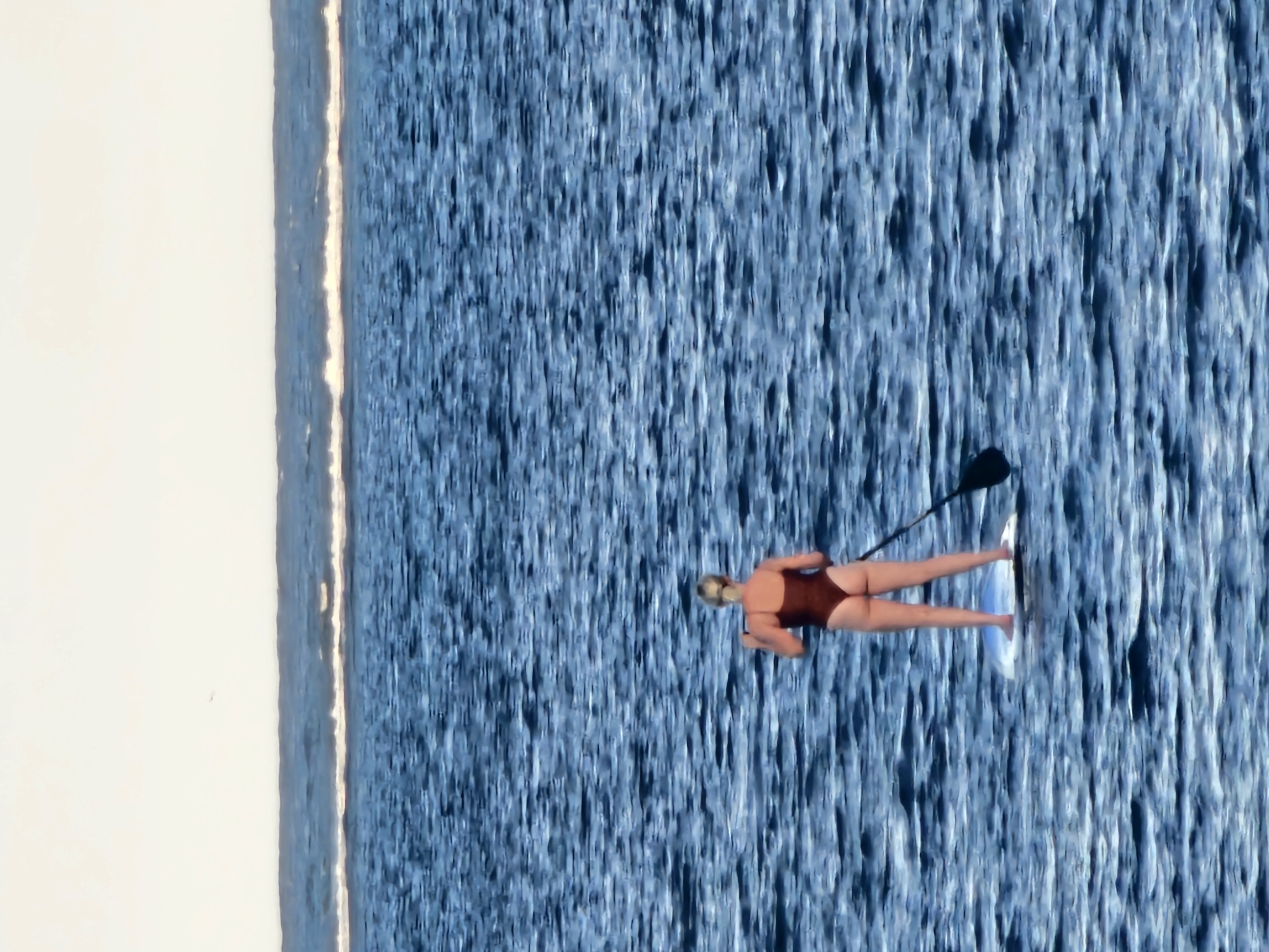 A woman with blonde hair, wearing a maroon one-piece swimsuit, is paddleboarding on a body of water. She is standing on a white paddleboard, holding a black paddle. The water is a deep blue with ripples and waves. In the distance, there is a faint line of white water, possibly a breaking wave or a reef. Above the water, the sky is a pale, hazy white. The scene appears to be set at sea, likely off the coast of Denpasar, Indonesia, given the tropical appearance of the water and sky.