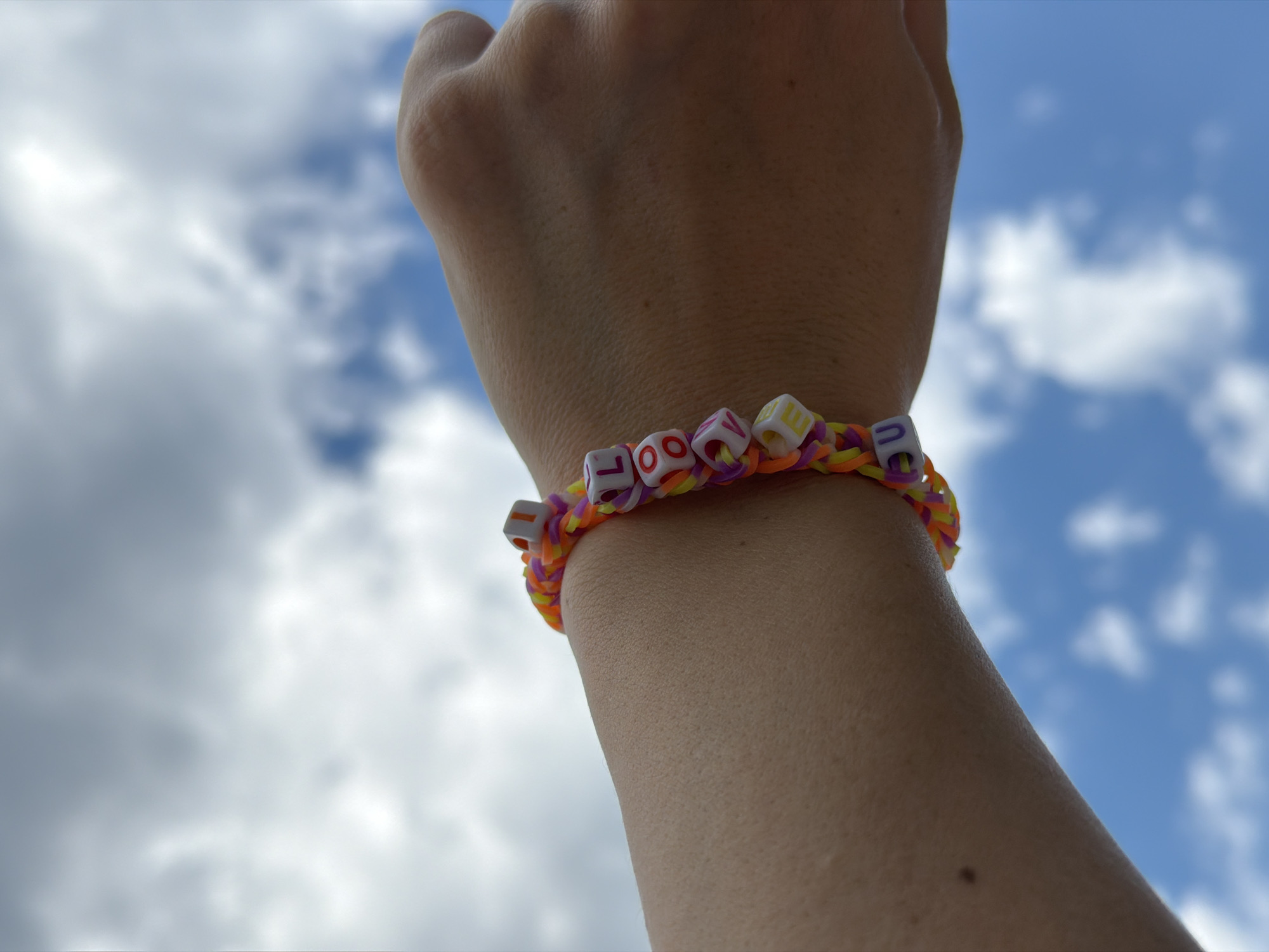 A person's hand and wrist are positioned in the foreground against a blurred background of a blue sky with scattered white clouds. The person is wearing a colorful loom band bracelet made of interlocking rubber bands in hues of orange, yellow, and purple. The bracelet is adorned with white cube-shaped beads with letters spelling out the word "LOVE". No specific details suggest the location is in Budva, Montenegro.