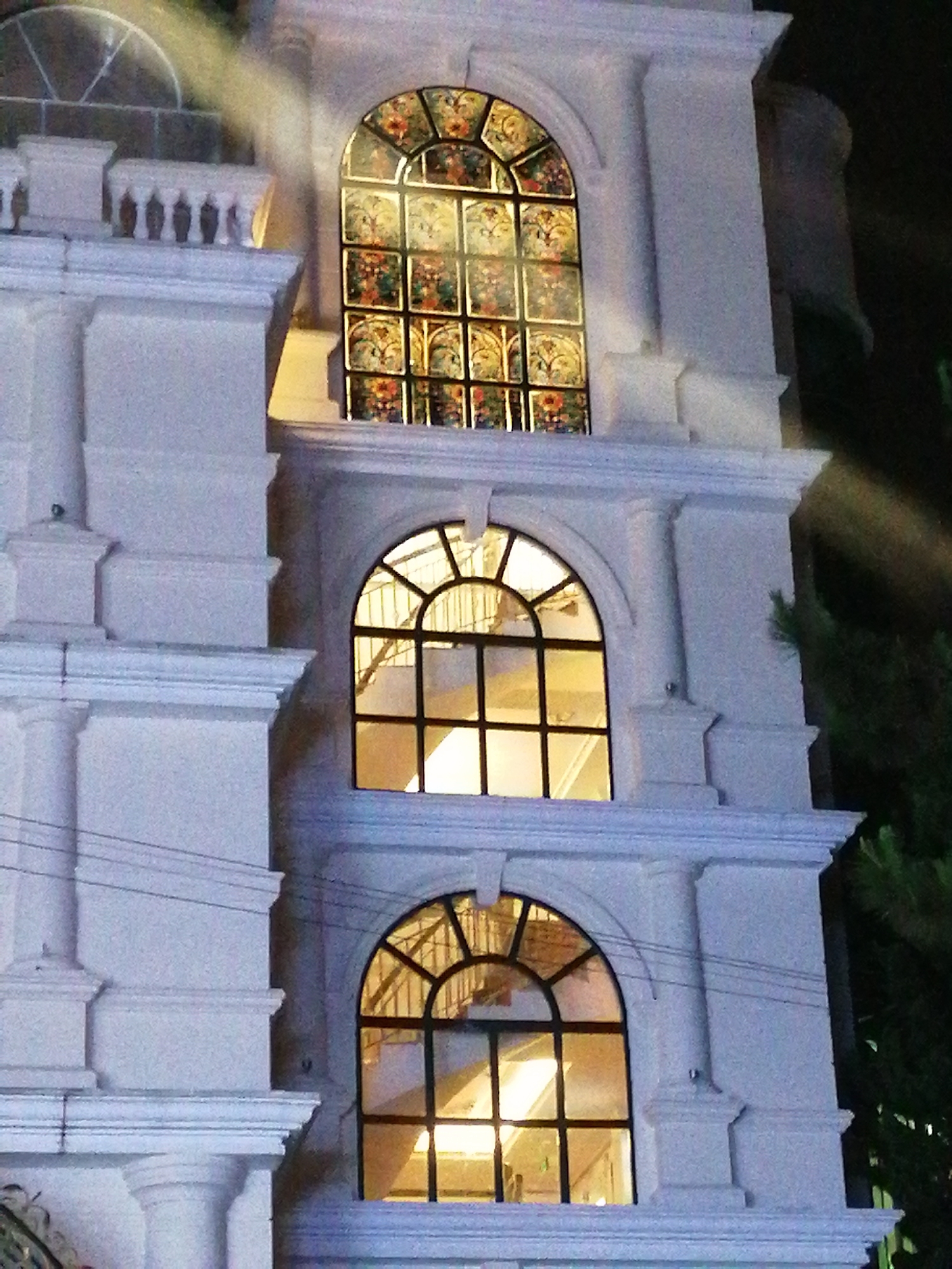 A nighttime photograph of a multi-storied, whitewashed architectural facade in Da Lat, Vietnam, displays a series of arched windows. The top window is stained glass, while the two beneath it are clear with black mullions. The building is detailed with classical architectural elements such as cornices and engaged columns. Part of a treetop appears on the right side. Overhead electrical wires run across the facade.