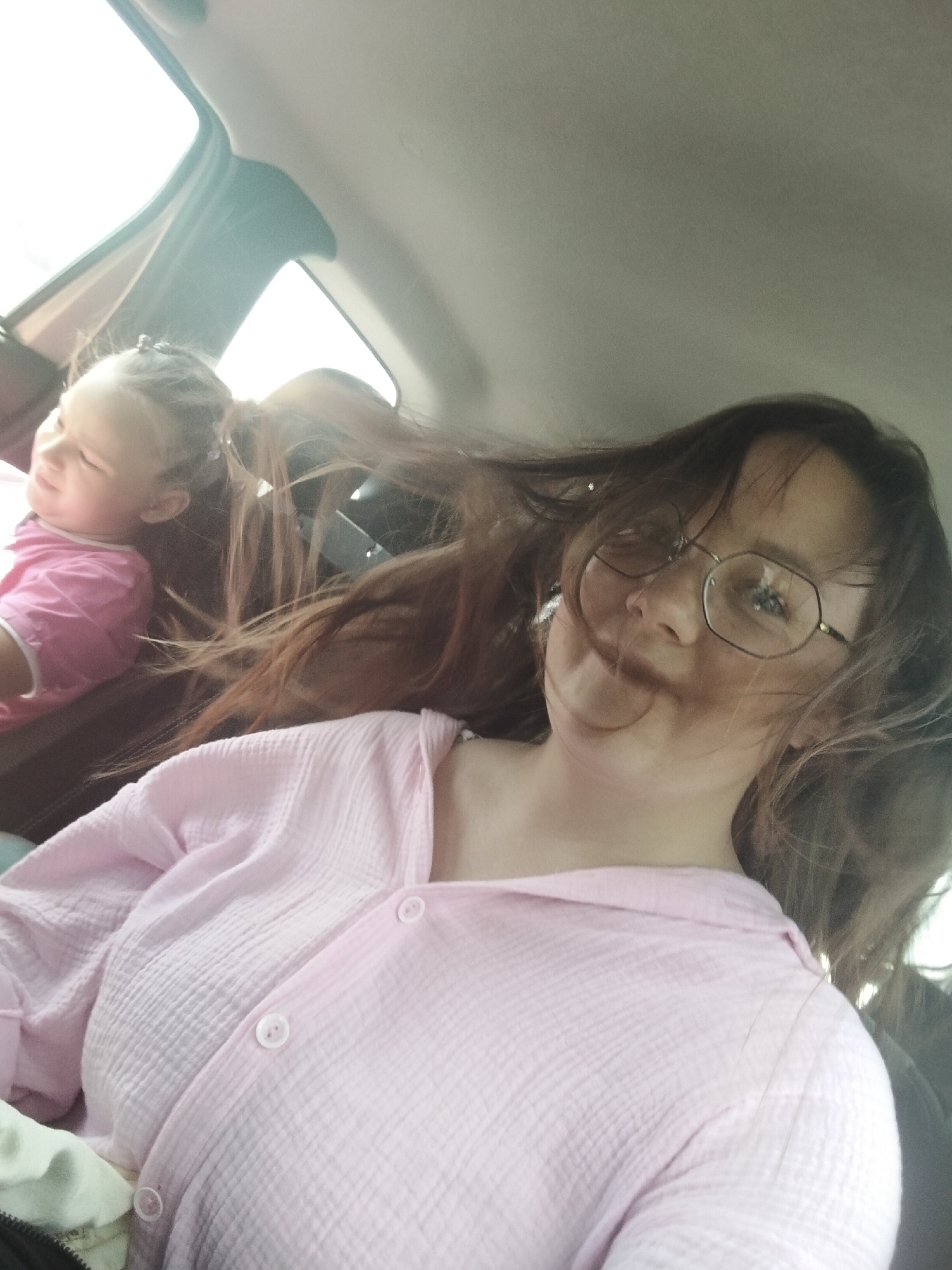 In a car interior, two females are visible. In the foreground, a woman with glasses and long brown hair is looking at the camera, wearing a pink collared shirt. Her hair is blowing in the wind. In the background, to the left, a young girl with blonde hair in a ponytail, wearing a pink shirt, is looking away from the camera. The car's interior is visible, with windows and seats. The image is an informal selfie, likely taken while traveling. No specific location in Блохино, Russia is identifiable.