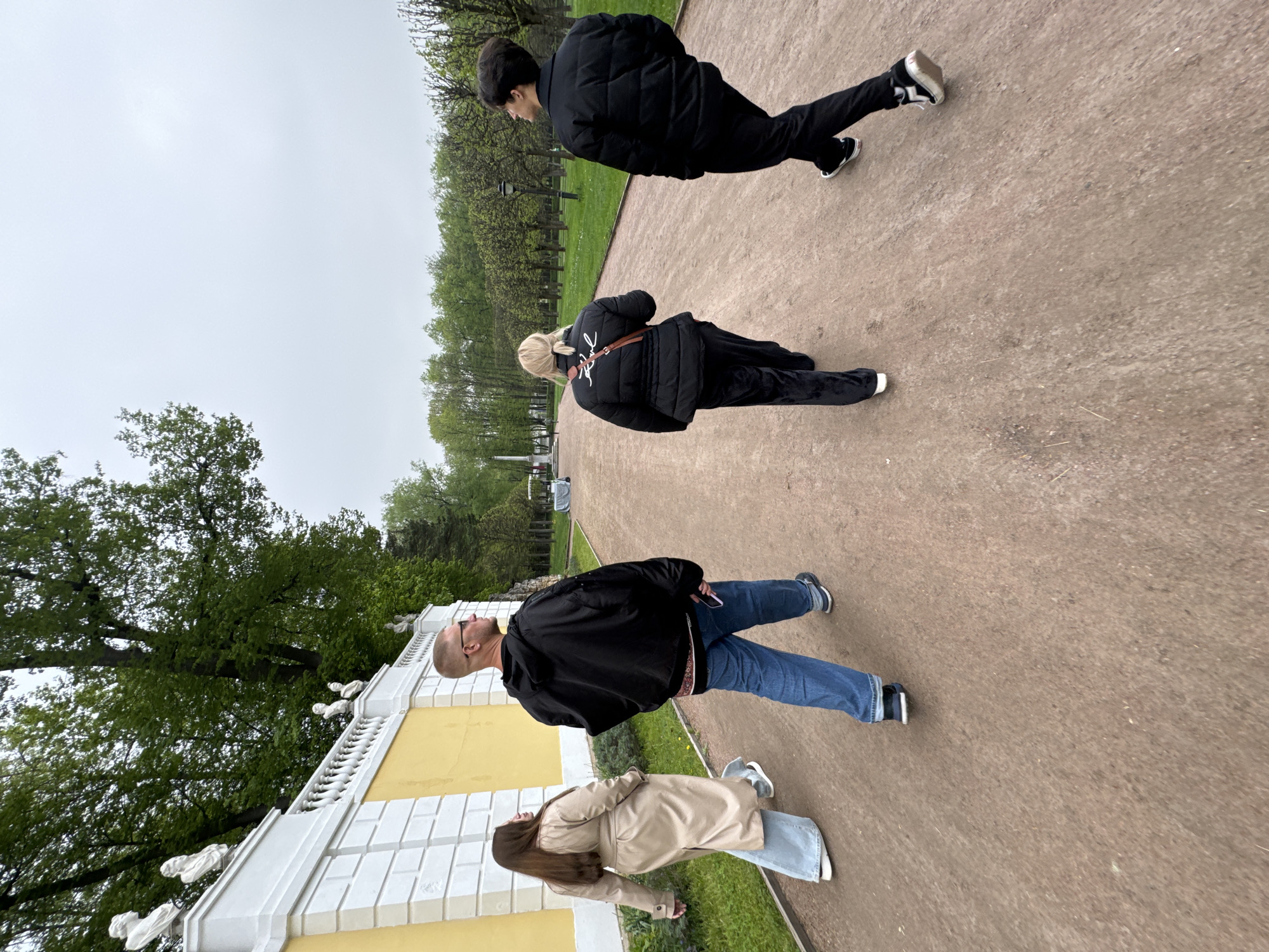 Four individuals are observed on a pathway within the State Museum Estate "Arkhangel'skoe" in Russia. The pathway, which is a reddish-brown color, appears to be lined with green grass on either side. The first individual is male, dressed in a black puffer jacket and black pants, and is walking away from the camera. The second individual, a female with blonde hair, is wearing a black puffer jacket with a brown strap and black pants and is also walking away from the camera. The third individual, a bald male wearing glasses, is wearing a black jacket and blue jeans. The fourth individual, a female with long brown hair, is wearing a tan trench coat and light blue jeans. A yellow and white wall is visible to the left, with trees lining the background. The sky appears overcast.
