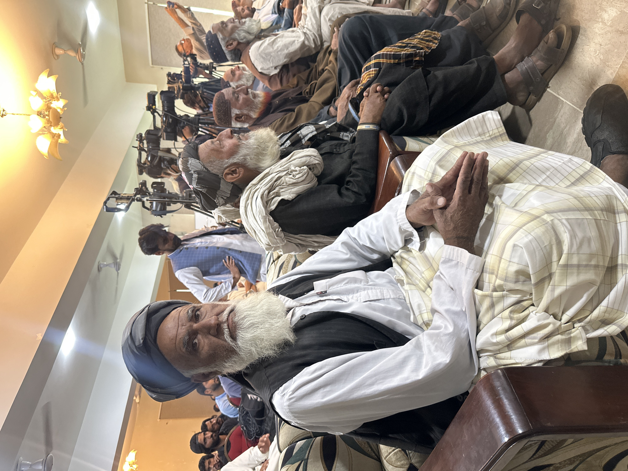 In Quetta City Tehsil, Pakistan, an indoor gathering shows a row of older men with white beards seated and facing forward, some wearing turbans and traditional clothing. At least one individual has their hands clasped. Behind them, multiple television cameras on tripods are positioned, suggesting a media event. Additional people are present in the background, visible in the lower part of the frame. A chandelier hangs from the ceiling, and the room is brightly lit. The footwear of some seated men includes sandals.