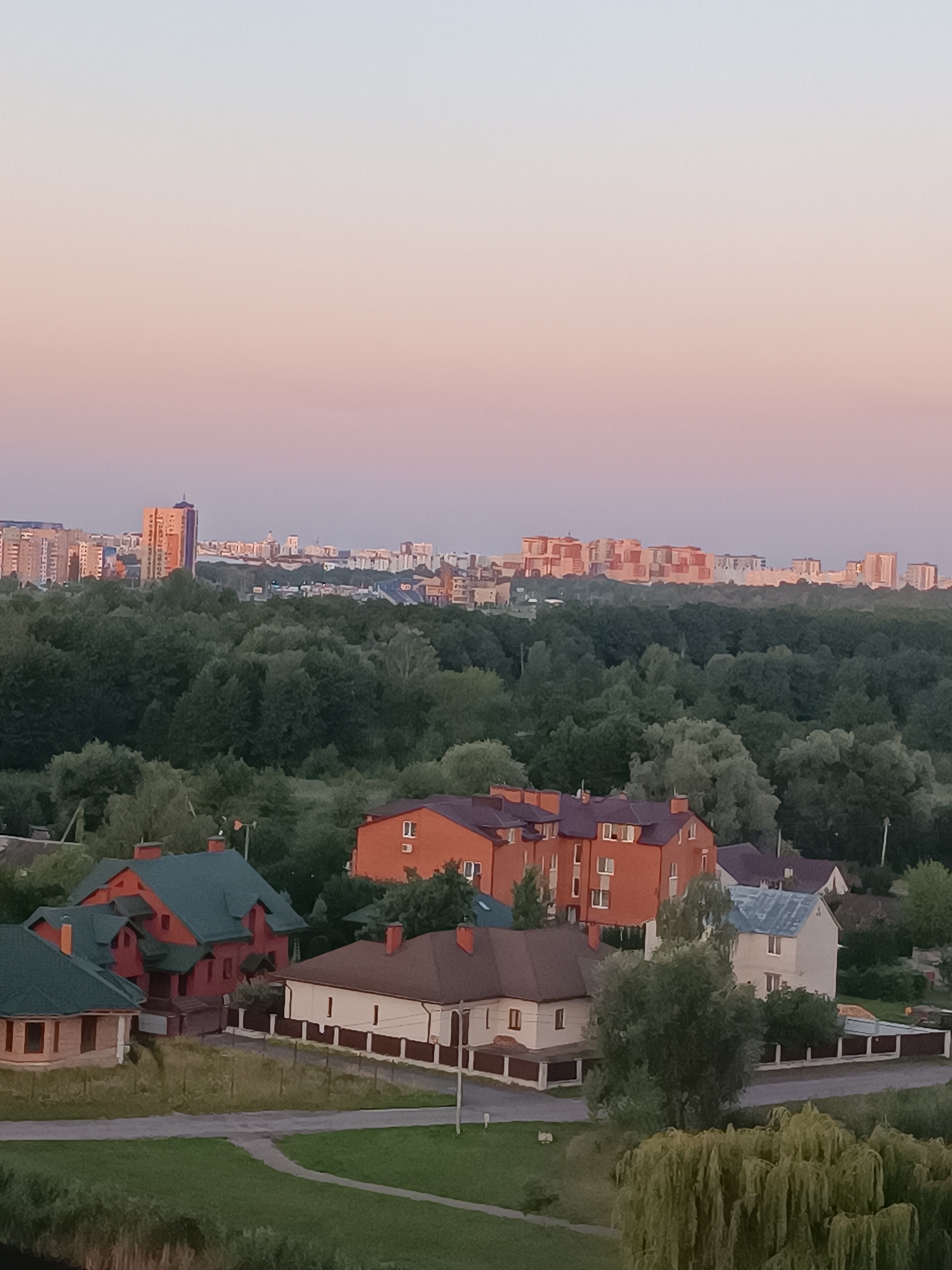 This image captures a panoramic view of a suburban landscape at dusk, with a cityscape visible in the background. In the foreground, several residential houses with varying roof colors and architectural styles are present, some featuring prominent chimneys. A winding road or path runs through the scene, bordered by green grass and trees. The middle ground is dominated by a dense forest, its dark green foliage contrasting with the softer hues of the sky. The distant horizon is occupied by a city skyline, characterized by a collection of multi-story buildings, including a distinctive tall, orange-colored tower. The sky exhibits a gradient of colors, transitioning from a pale blue at the top to soft pinks and purples near the horizon, indicative of a sunset. The overall atmosphere is serene and tranquil.