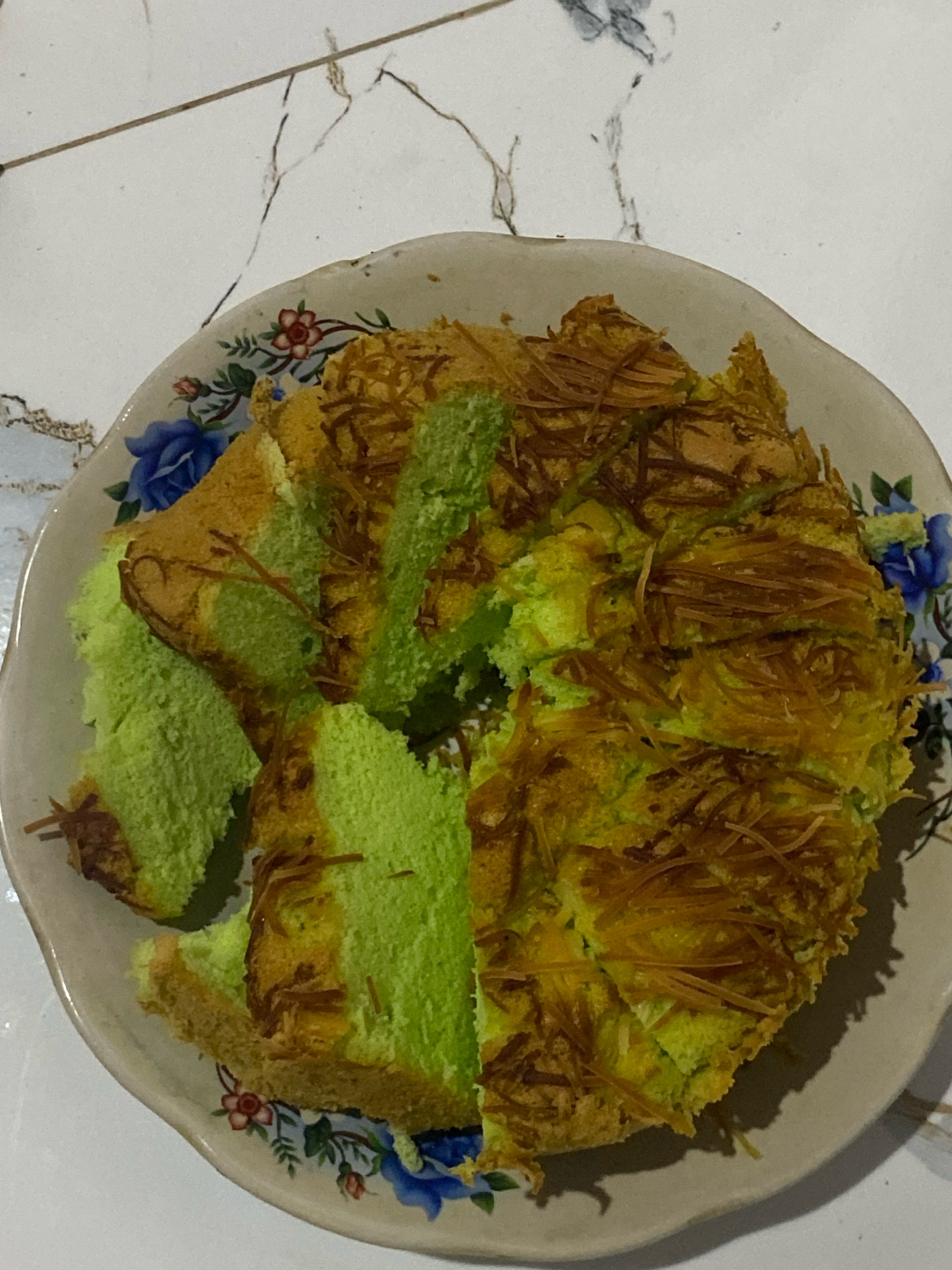 A sliced cake, with green sponge and grated cheese topping, rests on a decorative ceramic plate. The plate is adorned with blue and red floral patterns. The cake is presented on a white tiled surface, possibly a table or countertop. The lighting casts shadows on the cake and plate. The scene appears to be indoors, with no visible windows or outdoor elements. The specific location within Planjan, Indonesia, is not discernible from the image.
