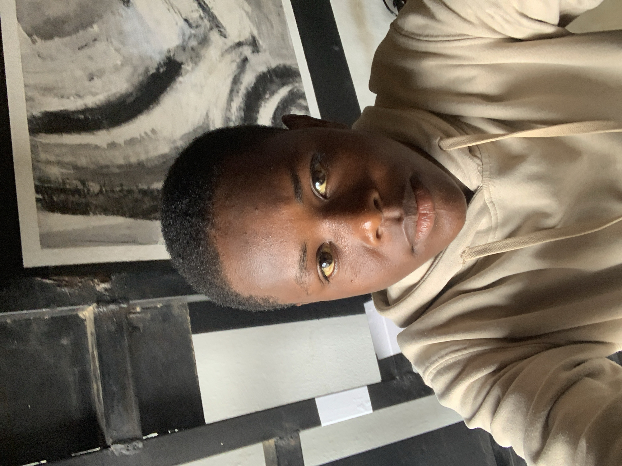 An indoor, close-up, portrait-style photograph, rotated 90 degrees counter-clockwise, features a young person, likely male, with dark skin and short, dark hair. The subject is looking directly at the camera with a neutral expression. Their eyes are a notable light brown or amber color. They are wearing a plain beige or light brown hooded sweatshirt, with the drawstrings of the hood visible around the neckline.
The background is an interior space. To the upper left, behind the subject's head, there is a framed piece of artwork or a poster featuring abstract, swirling patterns in shades of black, white, and gray. Below this, and partially visible in the lower right of the frame, are distinct black and white horizontal striped elements, possibly part of a wall, floor, or furniture. There are also darker, possibly wooden or metallic, structural components in the background. The lighting appears to be soft and even, suggesting indoor conditions. The setting offers no specific visual cues to its location beyond being an indoor space, but the context provided indicates it is Jos, Nigeria. No visible text is present in the image.