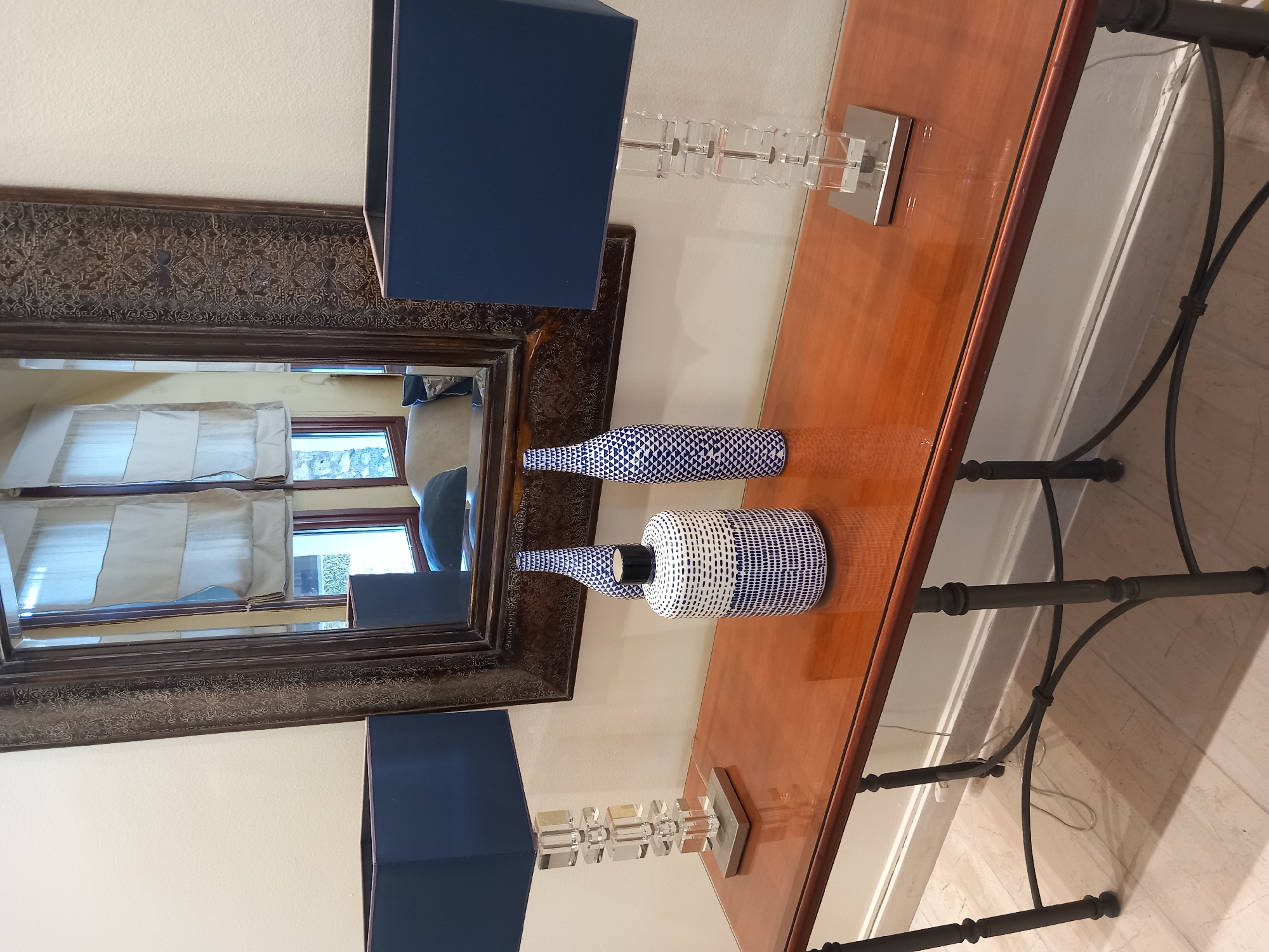 The image shows a wooden console table with a glass top, positioned against a white wall. Two identical blue lamps with square shades and clear acrylic bases are placed on either end of the table. In the center of the table, there are two decorative vases. One is a white vase with intricate blue patterns, and the other is a tall, slender blue and white patterned vase. A large, ornate mirror with a dark, patterned frame hangs on the wall behind the console table. The mirror reflects a window with draped curtains and a glimpse of a sofa with a blue cushion, suggesting an indoor setting. The floor is tiled with light-colored tiles. The overall scene depicts a stylishly decorated entryway or living space.