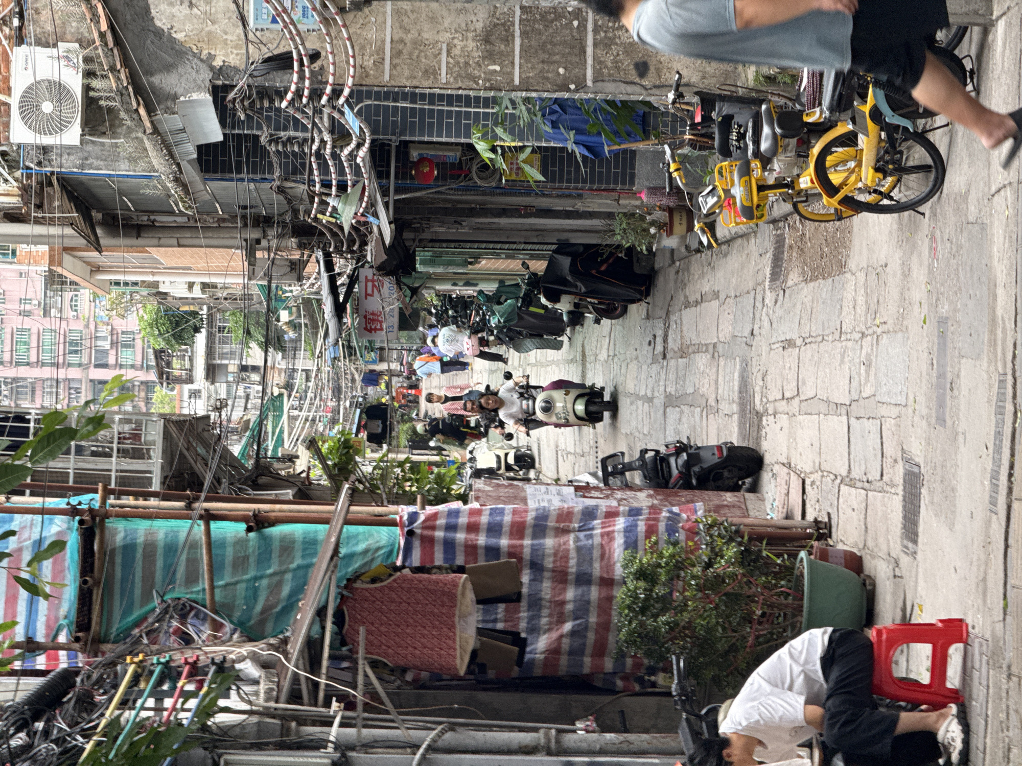 The image shows a narrow, congested alleyway in what appears to be Liwan District, China. The alley is paved with rectangular stone blocks. Along the alley's length, numerous parked motorbikes and scooters dominate the visual space, leaving limited room for passage. Overhead, a dense network of electrical wires and cables forms a chaotic web, typical of densely populated urban areas. Construction or repair work is evident, with scaffolding and protective tarpaulins obscuring building facades.
Several individuals are visible. One man in a gray shirt and dark shorts is walking along the alley. Another person is crouched low to the ground and is sitting on a red plastic stool with their head very close to the pavement.
The buildings visible are of varied heights and construction styles, and are characterized by features such as air conditioning units. Laundry lines stretch across parts of the alley. The presence of potted plants adds a touch of greenery to the otherwise utilitarian scene.