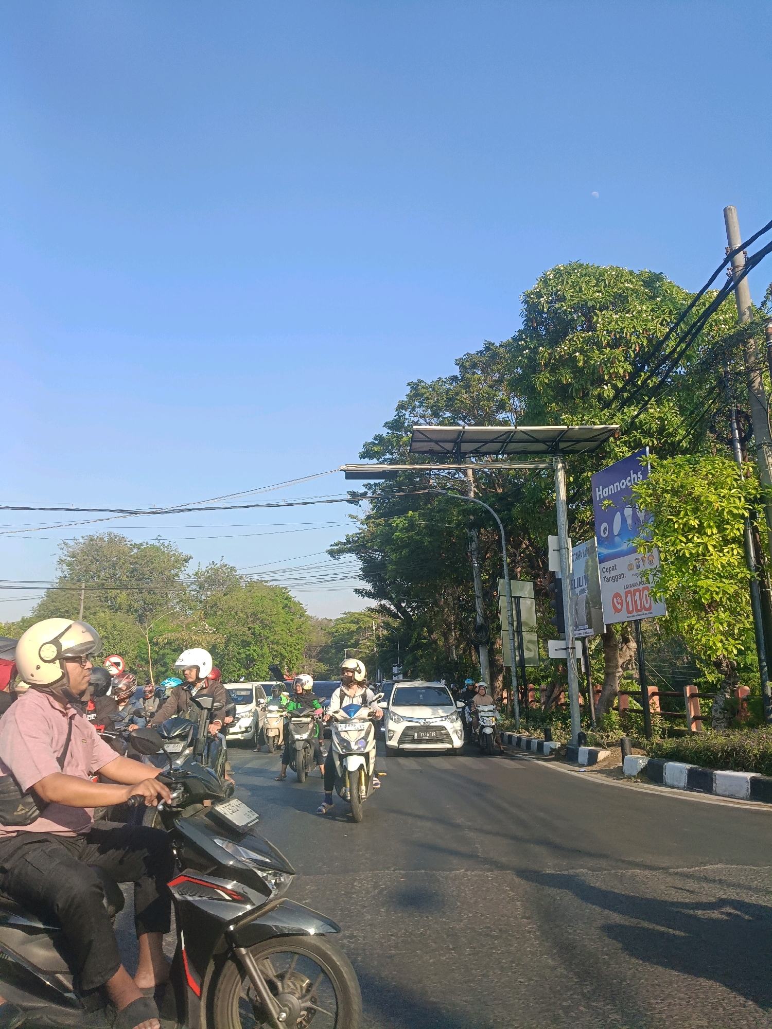 In a sun-drenched scene in Surabaya, Indonesia, a traffic jam unfolds on a paved road under a clear blue sky. Several individuals on motorcycles navigate the congestion. In the foreground, a person wearing a light pink shirt and black pants, with a light-colored helmet, rides a black motorcycle. To their right, other motorcyclists are visible, some wearing helmets. Behind the motorcycles, a white car and another white car are present. Lush green trees line the right side of the road, where a street lamp and a sign that reads "Hannochs" are positioned. The road has distinct black and white painted curbs. The image captures a typical urban traffic situation in Surabaya.