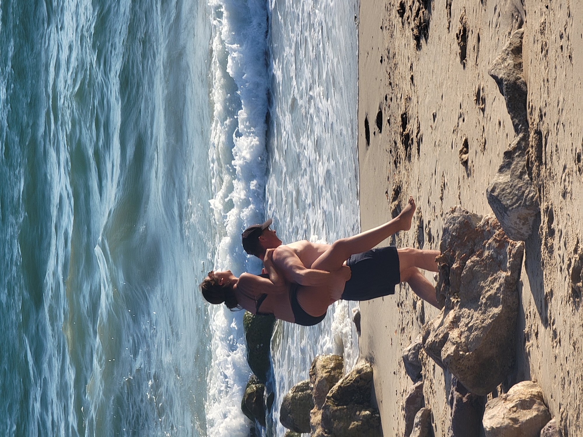 In Quarteira, Portugal, a man is giving a woman a piggyback ride on the beach. The man, wearing dark swim trunks and a black baseball cap, walks on the sand near the ocean. The woman, in a black bikini, sits on his shoulders, looking to her right. The ocean behind them has waves breaking on the shore and large rocks are scattered on the beach in the foreground.
