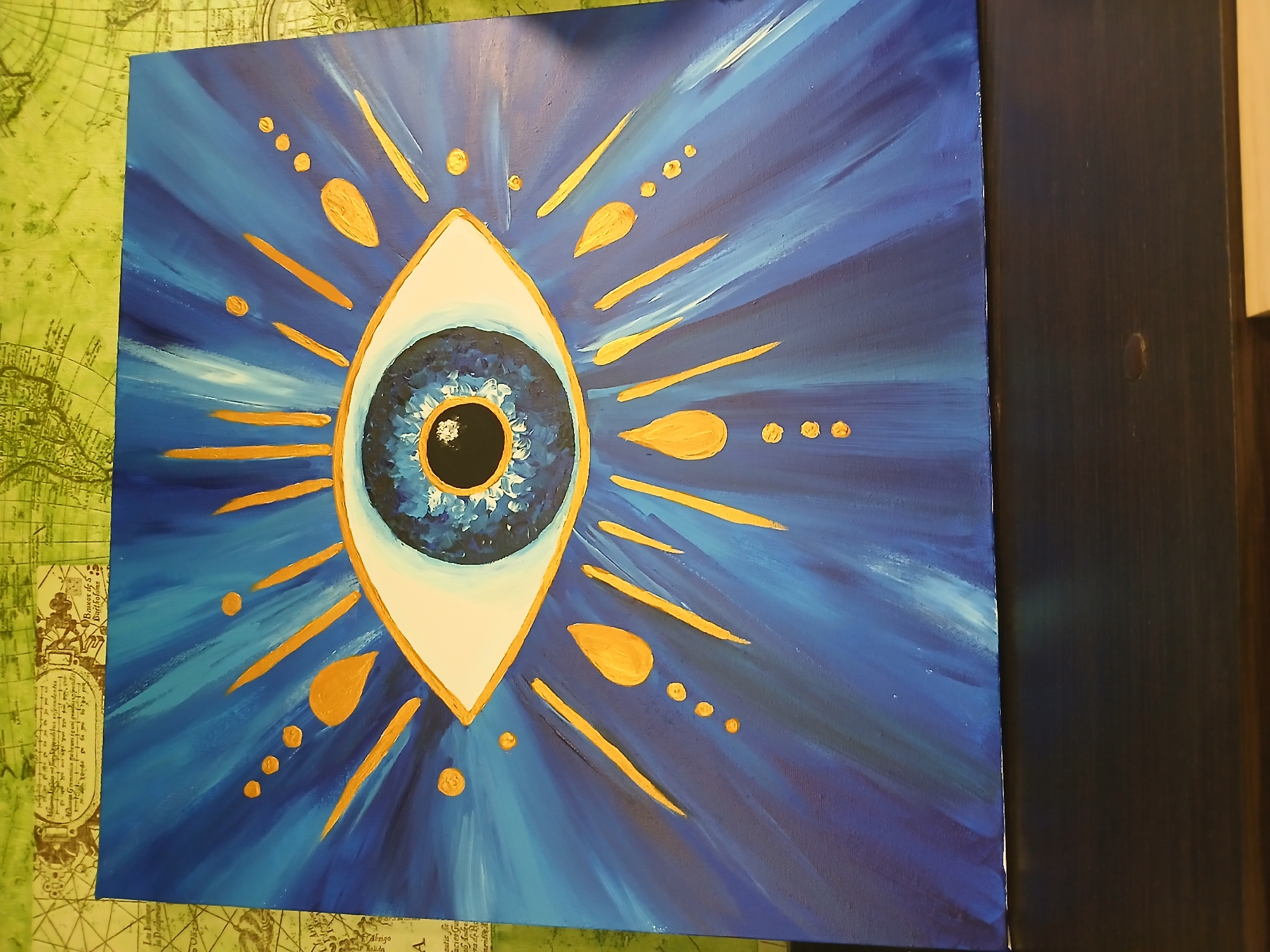 The image shows a square painting of a stylized blue eye with radiating gold designs, placed on top of a green surface that has a pattern resembling an old map. The eye is almond-shaped with a white border outlined in gold. Inside, the iris is a swirl of blues surrounding a black pupil. Radiating outwards from the eye are strokes of blue paint, suggesting light or energy. Interspersed among these strokes are golden lines and shapes, some like rays and others like teardrops, arranged symmetrically. The painting has a vibrant and symbolic quality, suggesting protection or awareness. The image does not provide any information to suggest the location is Kamianske, Ukraine.