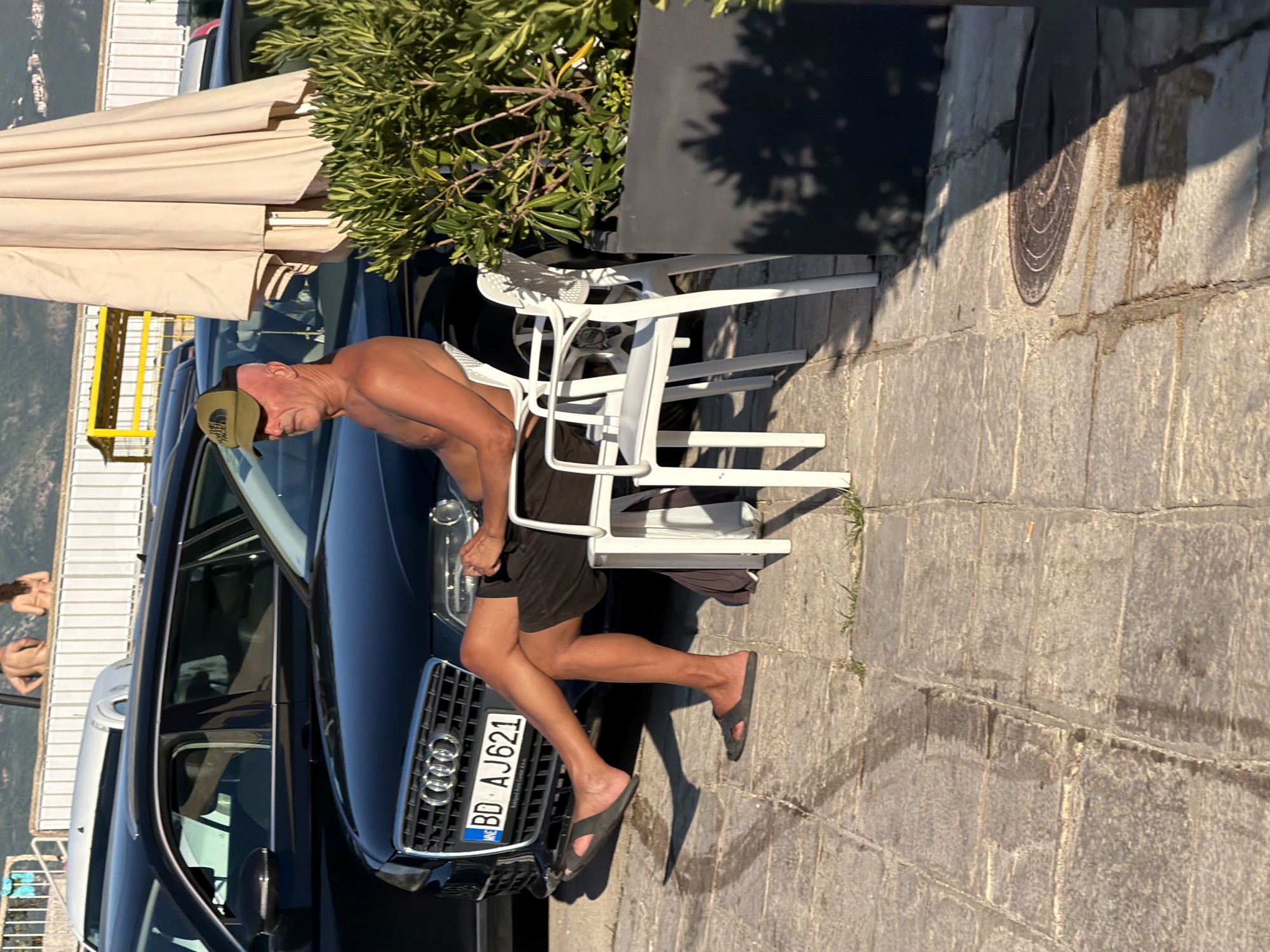 A man with a shaved head and a green cap is sitting on a white plastic chair facing right, with his legs crossed. He is wearing black shorts and is shirtless, with his skin appearing sunburned. He is seated on a paved outdoor area with large rectangular stone tiles. Behind him, a black Audi car is parked. To the right of the man, another white plastic chair is empty, and a large potted plant with green leaves is visible. In the background, there is a light-colored umbrella, a fence, and distant hills. The scene appears to be in Budva, Montenegro, possibly near a beach or promenade, given the presence of cars parked and the general atmosphere.