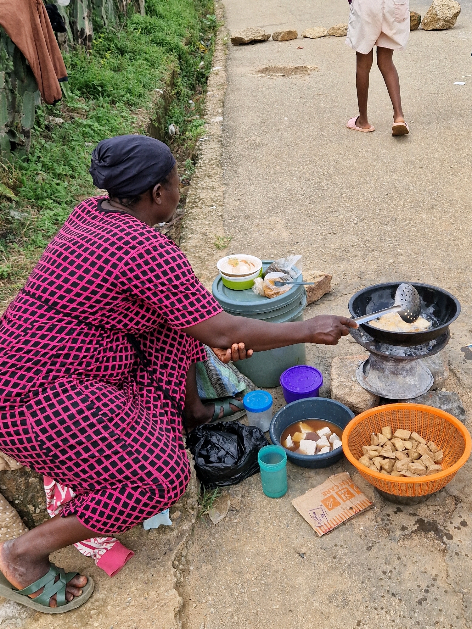 In an outdoor scene on a paved surface, a woman wearing a pink and black checkered dress and a dark head covering is kneeling, tending to a cooking area. She is holding a metal slotted spoon over a black pot on a makeshift stove composed of stacked stones and metal rings. Inside the pot, food is being fried in oil. Around her are various containers: a grey bucket with a green lid holding food, a blue bucket, a purple lidded container, a grey bowl with cut-up white food items submerged in a brown liquid, and an orange basket filled with cubed, light brown food. A blue plastic cup and a crumpled piece of cardboard are also on the ground. To the left of the woman, greenery and a brown garment hang from a cactus-like plant. In the background, a person wearing white shorts and pink sandals is walking away. The ground is a mixture of concrete and dirt, with scattered stones.