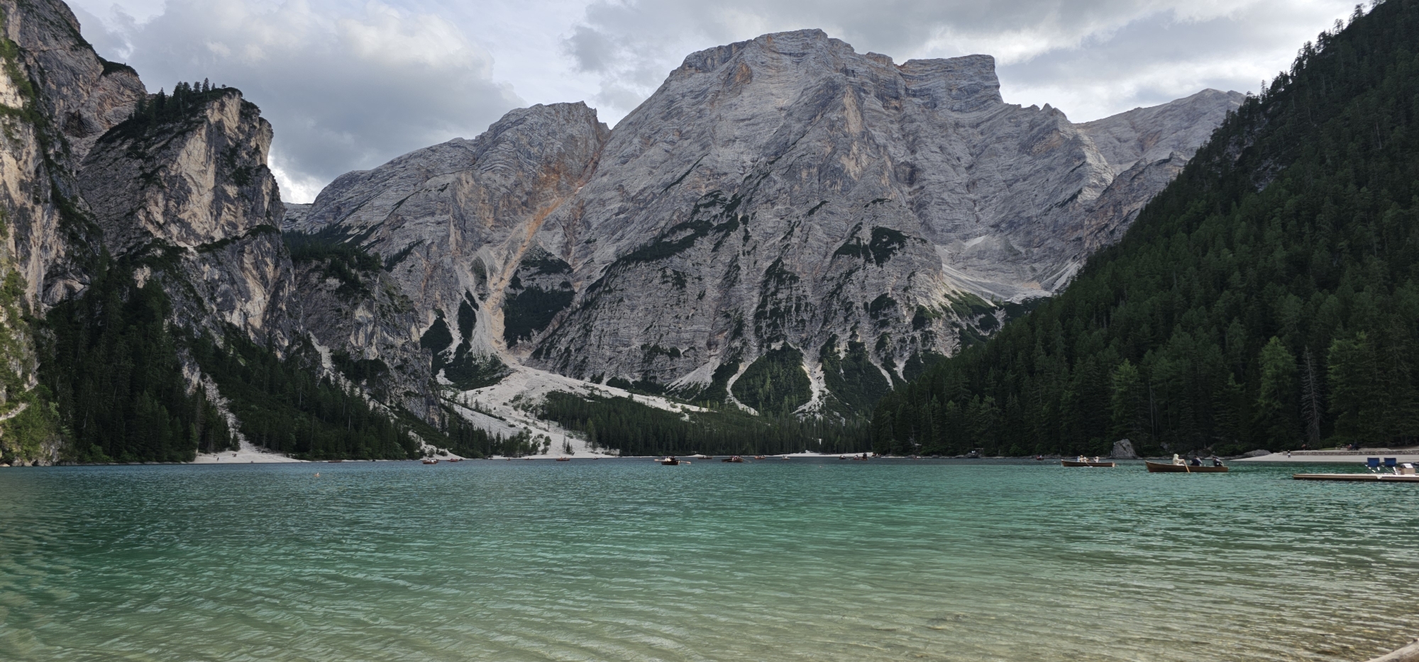 This is a wide-angle, landscape photograph of Lago di Braies (Pragser Wildsee) in the Prags Valley, South Tyrol, Italy. The foreground features clear, turquoise water with visible ripples, and a sandy or pebbly bottom near the shore. Several small, wooden boats are scattered across the lake. In the middle ground, a narrow, white beach is visible, bordered by a dense forest of dark green pine trees. Behind the trees, imposing, grey, rocky mountains rise steeply, with some areas of exposed rock and scree. The sky is overcast with soft, grey clouds. A few people are visible on the beach and in the boats, engaged in recreational activities.