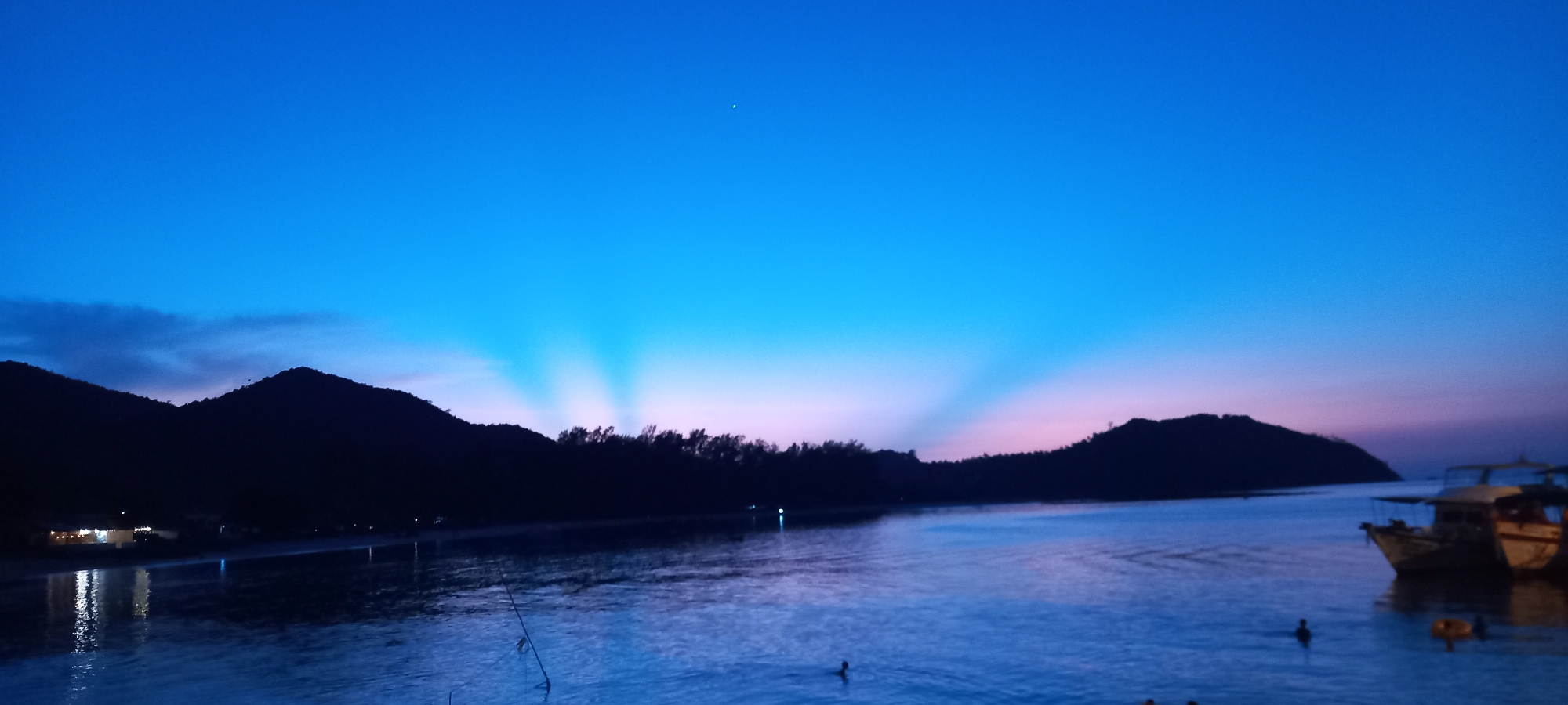 The image depicts a coastal scene in Baan Chaloklum, Thailand, at dusk. In the foreground, a body of water reflects the ambient light, appearing blue. Several individuals are seen swimming in the water. To the right, two boats are docked. In the mid-ground, the coastline is defined by a dark treeline and two elevated landmasses, one on the left and one on the right. The sky transitions from a light blue at the top to a pinkish-purple near the horizon, with crepuscular rays emanating upwards through gaps in the foliage. A bright spot, likely a star or planet, is visible in the upper center of the sky. A faint shoreline with building lights is visible to the far left. A fishing pole extends into the water on the left.