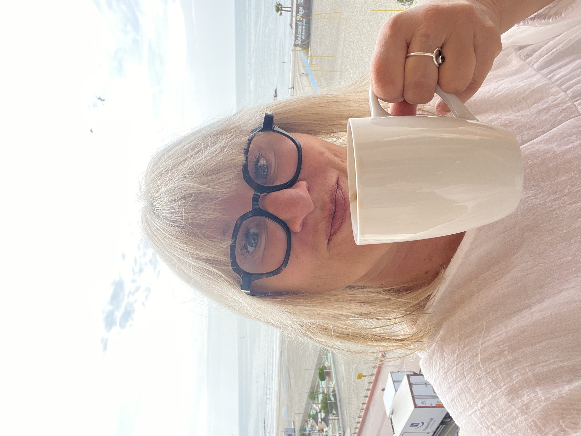 This media file captures a close-up, rotated portrait of a woman looking towards the viewer, with a bright coastal scene as her backdrop.
The main subject is a woman with blond, shoulder-length hair, wearing black-framed glasses and a light-colored, possibly white, textured top. She is holding a plain white ceramic mug with her right hand, on which she wears a silver-colored ring with a dark stone on her ring finger. Her eyes are visible through her glasses, and her lips are slightly parted.
The setting appears to be a balcony or elevated position overlooking a sandy beach and the ocean. In the background, a wide expanse of light-colored sand stretches towards the calm, light blue ocean. Along the beachfront, structures are visible, including what appears to be a beach restaurant with a sign that clearly reads "Restaurant Plage." Two palm trees stand beside this structure. Other beach amenities like yellow poles (possibly for umbrellas or demarcation) and blue objects (perhaps chairs or loungers) are discernible. Further down the beach, white containers or small vehicles are present, one of which shows the partially visible text "Van Oosten."
The sky is bright and appears to be daytime, with some soft clouds. Two small bird silhouettes are seen in the upper left portion of the sky, indicating fair weather. The overall location context is Palavas-les-Flots, France, consistent with a European coastal resort town. The image conveys a relaxed atmosphere, possibly enjoying a beverage with a view.