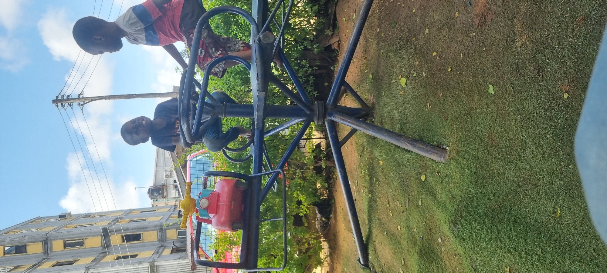 Two young Black boys are on a playground merry-go-round in Ikoyi, Nigeria. The boy on the left is wearing a dark t-shirt and blue jeans, and is sitting on a red plastic vehicle that is part of the merry-go-round. The boy on the right is wearing a red, white, and blue striped shirt and floral patterned shorts. He is sitting on the merry-go-round with his legs dangling. The merry-go-round is blue and has metal bars. Behind the merry-go-round are green bushes and a red vehicle. In the background, there is a tall, multi-story building with yellow and gray accents, and a utility pole with wires. The sky is blue with white clouds. The ground is covered in green grass and some fallen leaves.