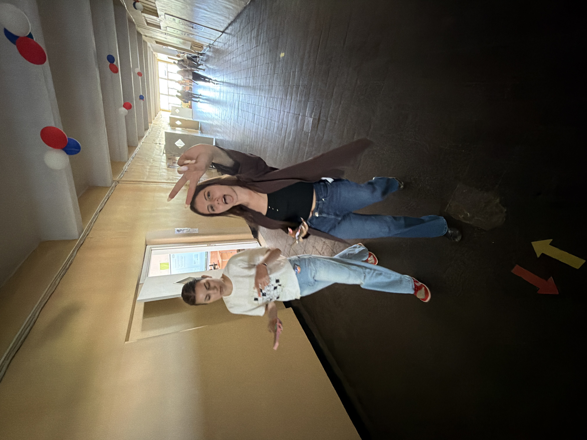 Two young women pose in a corridor. The corridor has red, white, and blue balloons attached to the ceiling, and painted directional arrows on the floor. The corridor has walls that are yellow on the left and brown on the right, and there are multiple people visible in the distance. The woman closer to the camera is wearing a white t-shirt, jeans, and red shoes. The other woman is wearing a brown jacket, black top, and jeans. It is likely that the location is in the city of Burgas, Bulgaria.