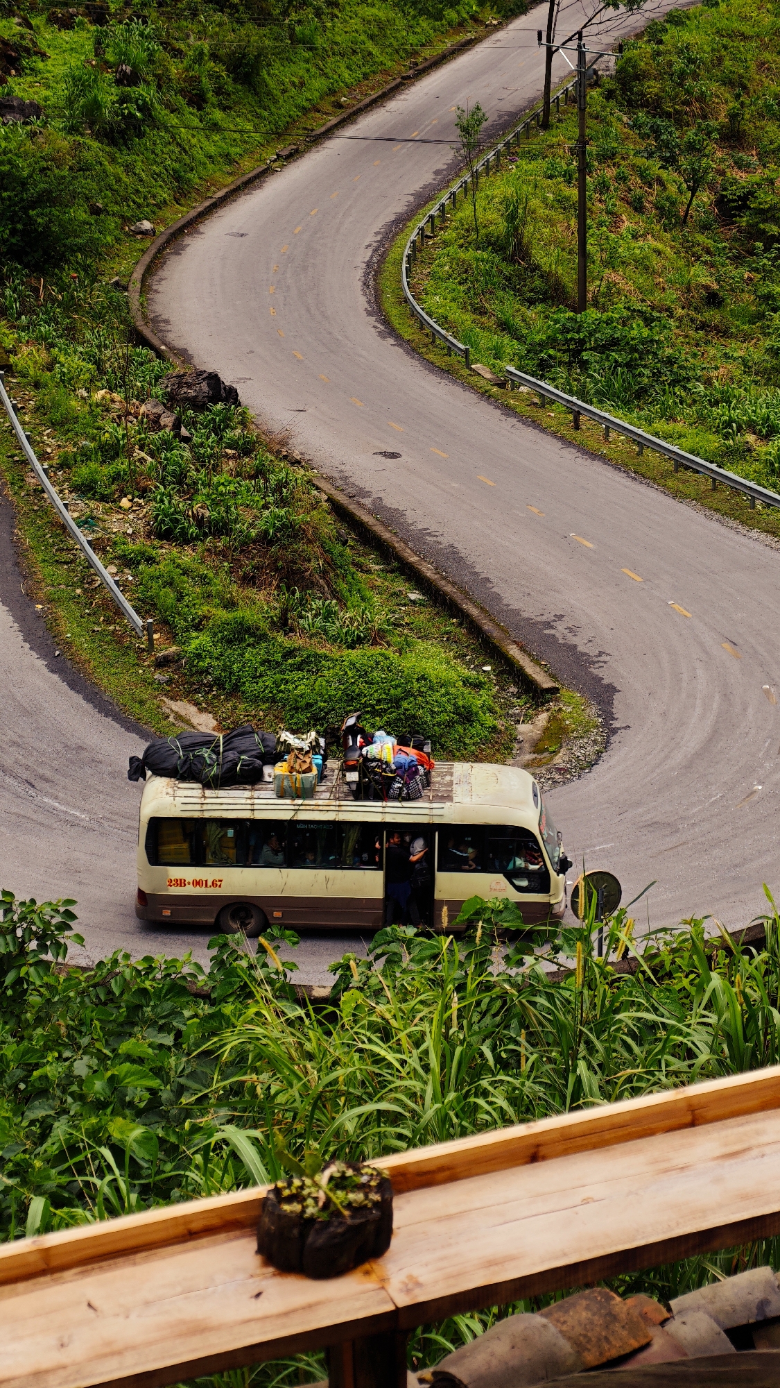 A medium shot photograph shows a winding road in Phố Bảng Commune, Vietnam. A beige passenger van, its roof laden with luggage and a motorcycle, is paused on the road. The van is marked with the number "23B-001.67". A person stands in the open doorway of the van. The road winds through green hills, and a metal guardrail runs along part of it. In the foreground, a wooden surface with a plant pot is visible, suggesting the photo was taken from an elevated position.