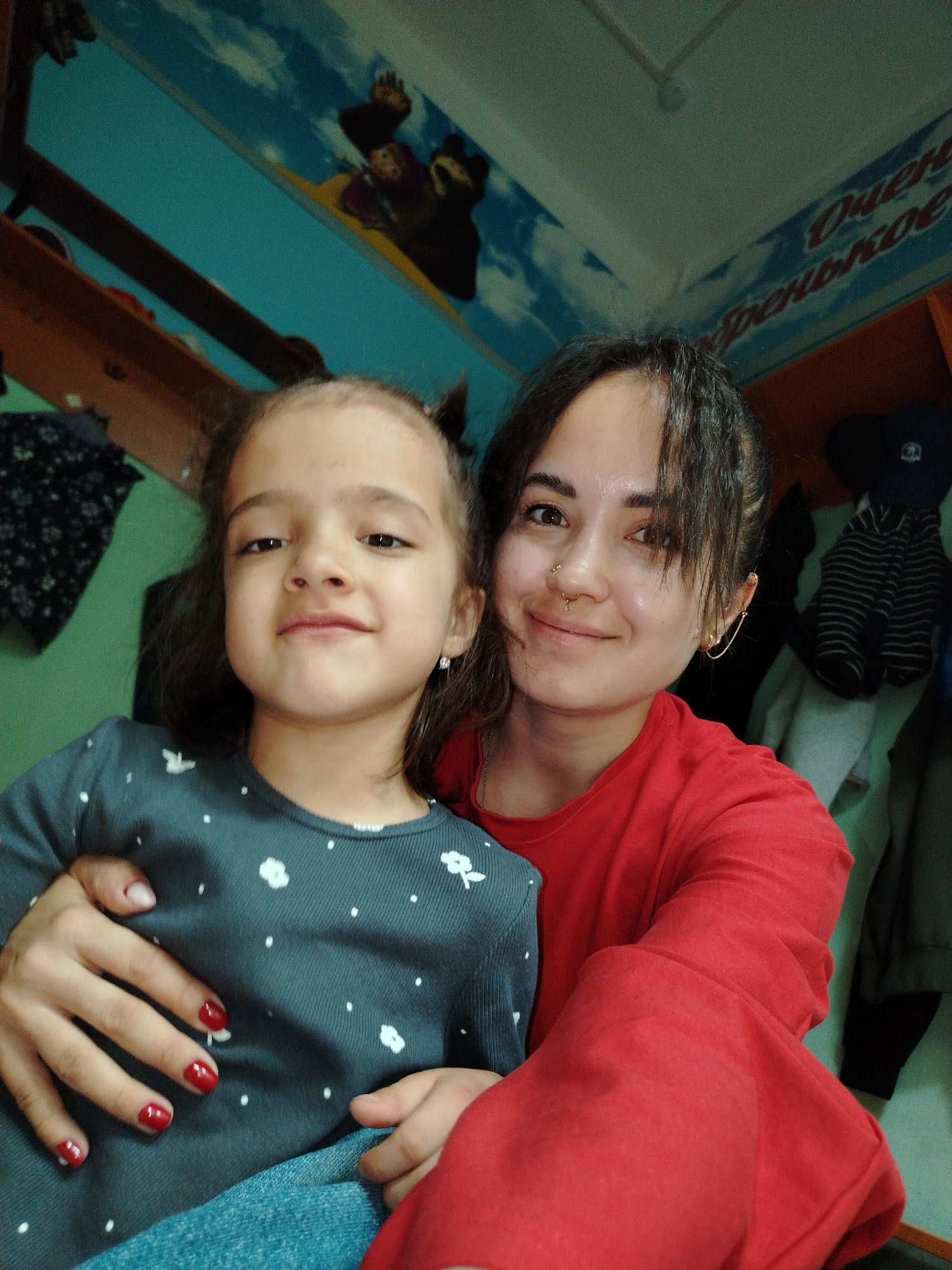 In the city of Chaikovsky, Russia, a medium shot captures a woman and a young girl, possibly mother and daughter, posing for a selfie indoors. The woman, on the right, is wearing a red top and has dark hair pulled back in a bun. She has a nose ring and an earring in her upper left ear. She is embracing the girl, who is on the left and wearing a dark gray long-sleeved shirt with white polka dots. The girl has dark hair and is looking towards the camera with a slight smile. Both individuals appear to be in good health. The background shows a blue wall with a mural of cartoon bears and clouds, suggesting a child's room or play area. Clothing items hang on hooks on the right side of the frame, and a wooden shelf is visible above. The lighting is somewhat dim, creating a soft ambiance. The overall scene conveys a sense of intimacy and family connection.