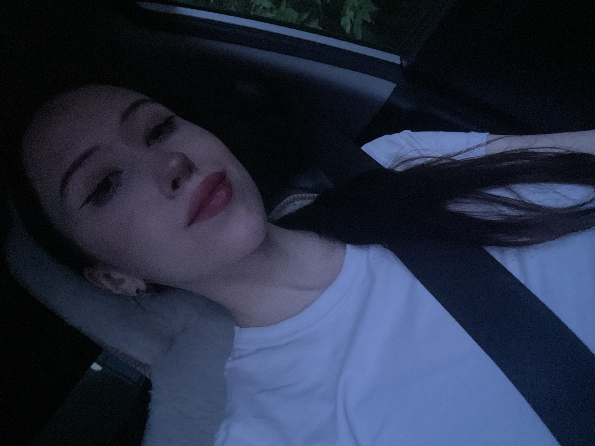A young woman with dark hair is seen from the chest up, sitting in a vehicle. She is wearing a white t-shirt and a black seatbelt. Her hair is pulled back into a ponytail and falls over her shoulder. The woman is wearing winged eyeliner and pink lipstick. She is looking slightly down and to her left, with her eyes partially closed. The lighting suggests it is either dusk or nighttime, as the surroundings are dark. The background is out of focus, but hints of greenery can be seen through a window on the right side of the frame, suggesting an outdoor environment. The overall mood of the image is subdued and intimate. No specific location in Votkinsk, Russia can be identified from the image.