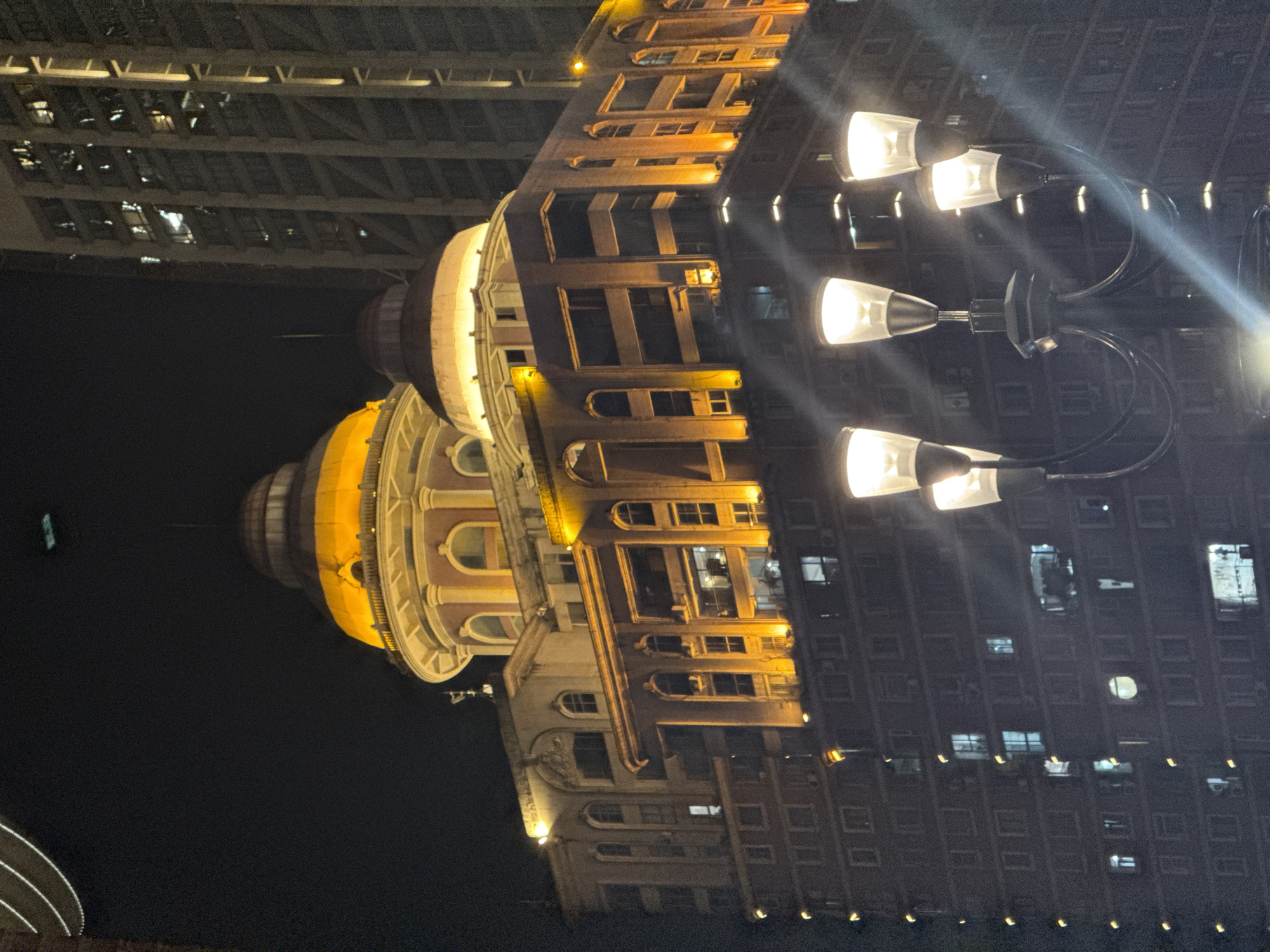 An outdoor, nighttime, medium-shot photograph captures the illuminated upper floors of a multi-story building in the Yuzhong District, China. The building features a prominent, glowing orange dome and a lighter colored dome, situated above a section with arched windows and vertical illumination. Below this, another section of the building displays numerous windows, some lit from within, and a facade lit with orange lights. In the foreground, a dark lamppost with multiple bright lights obscures portions of the building. A tall, modern skyscraper with vertical lighting is visible to the right of the dome-topped building, and a dark, slightly curved object is partially visible at the top left of the frame. The sky is dark and appears to be night.