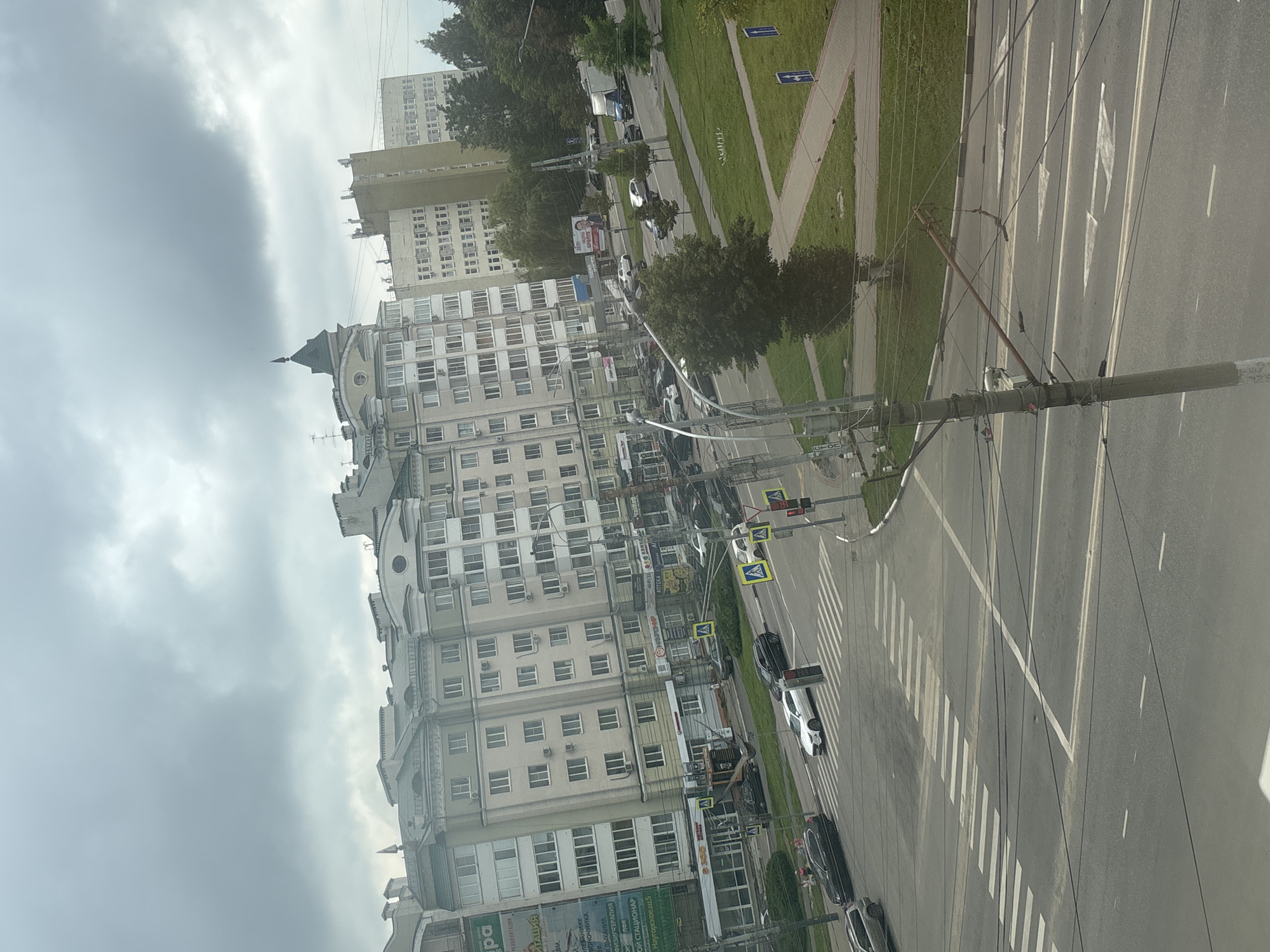 The image depicts an urban scene with a street and buildings, likely taken from an elevated viewpoint. The sky is overcast with heavy clouds. In the foreground, a multi-lane road with crosswalks is visible, with several cars driving and parked. Pedestrian crossings are marked with white stripes. Traffic lights are present at an intersection. Adjacent to the road, there are sidewalks and grassy areas with trees and some bushes. Several multi-story residential and commercial buildings line the street. One building is particularly tall and has a distinctive green, pointed turret. Power lines and utility poles are prominent in the scene. A few people are visible walking on the sidewalk. The overall impression is of a moderately busy urban environment on a cloudy day. The location is identified as Trosnikovo, Russia.