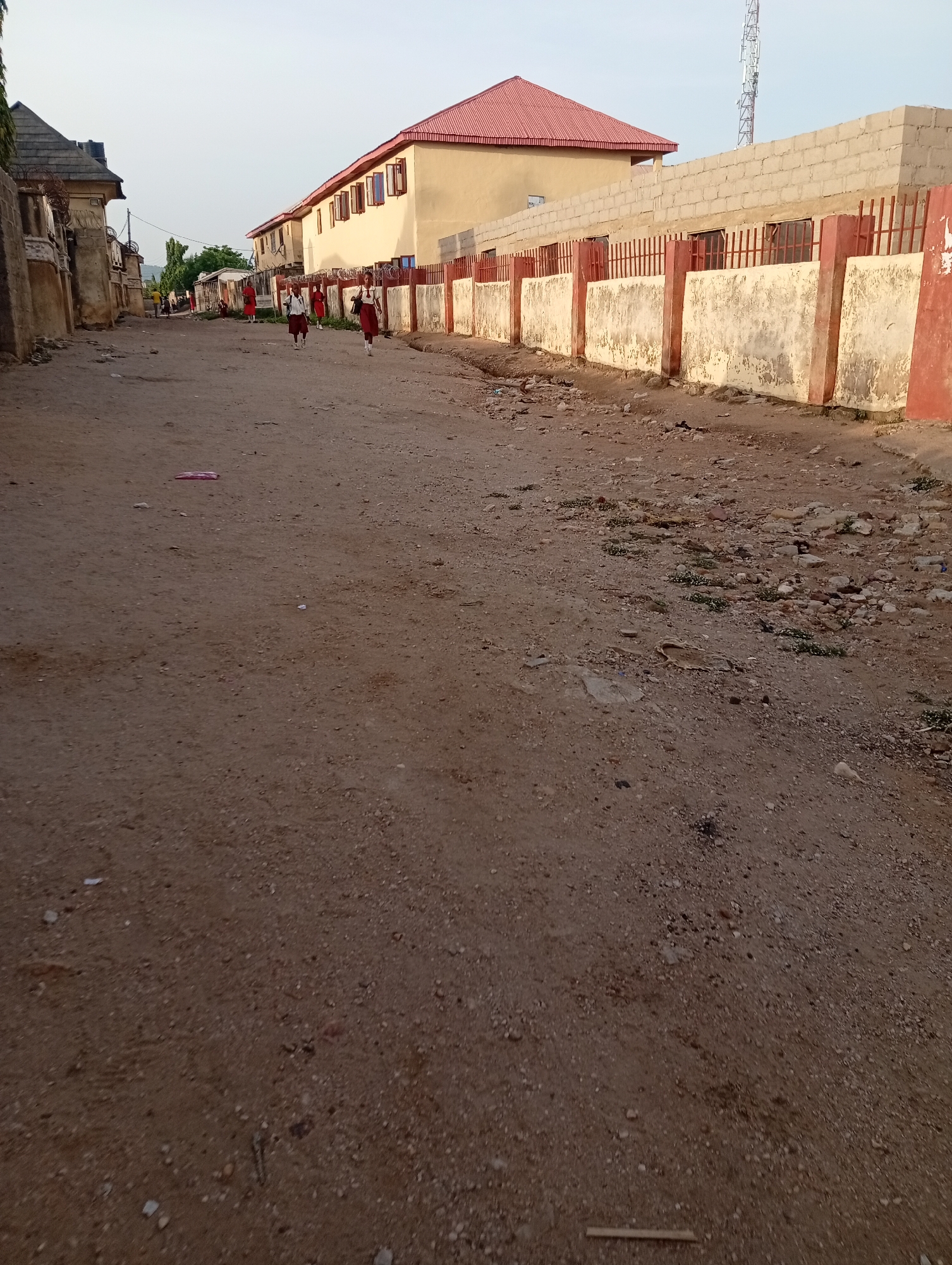 A daytime exterior shot reveals a dirt road in Adankolo, Nigeria, flanked by buildings and a wall. Three schoolgirls in red and white uniforms walk along the road. The building on the right has a red roof. A communication tower is visible behind the wall. Litter is scattered on the ground.