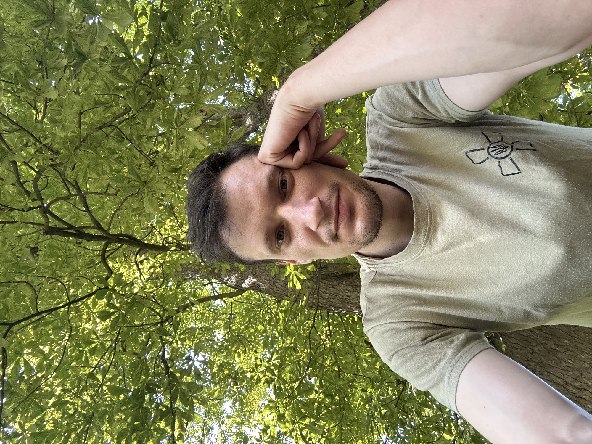 This is a selfie of a man with dark hair and a beard, resting his head on his hand. He is wearing a light olive green t-shirt with a black emblem on the left chest. The emblem is a stylized cross with a trident in the center. He is outdoors, with a large tree with green leaves in the background, suggesting a natural or park setting. The lighting indicates it is daytime, with sunlight filtering through the leaves. There is no visual information in the image that places the location in the city of Makiv, Ukraine.