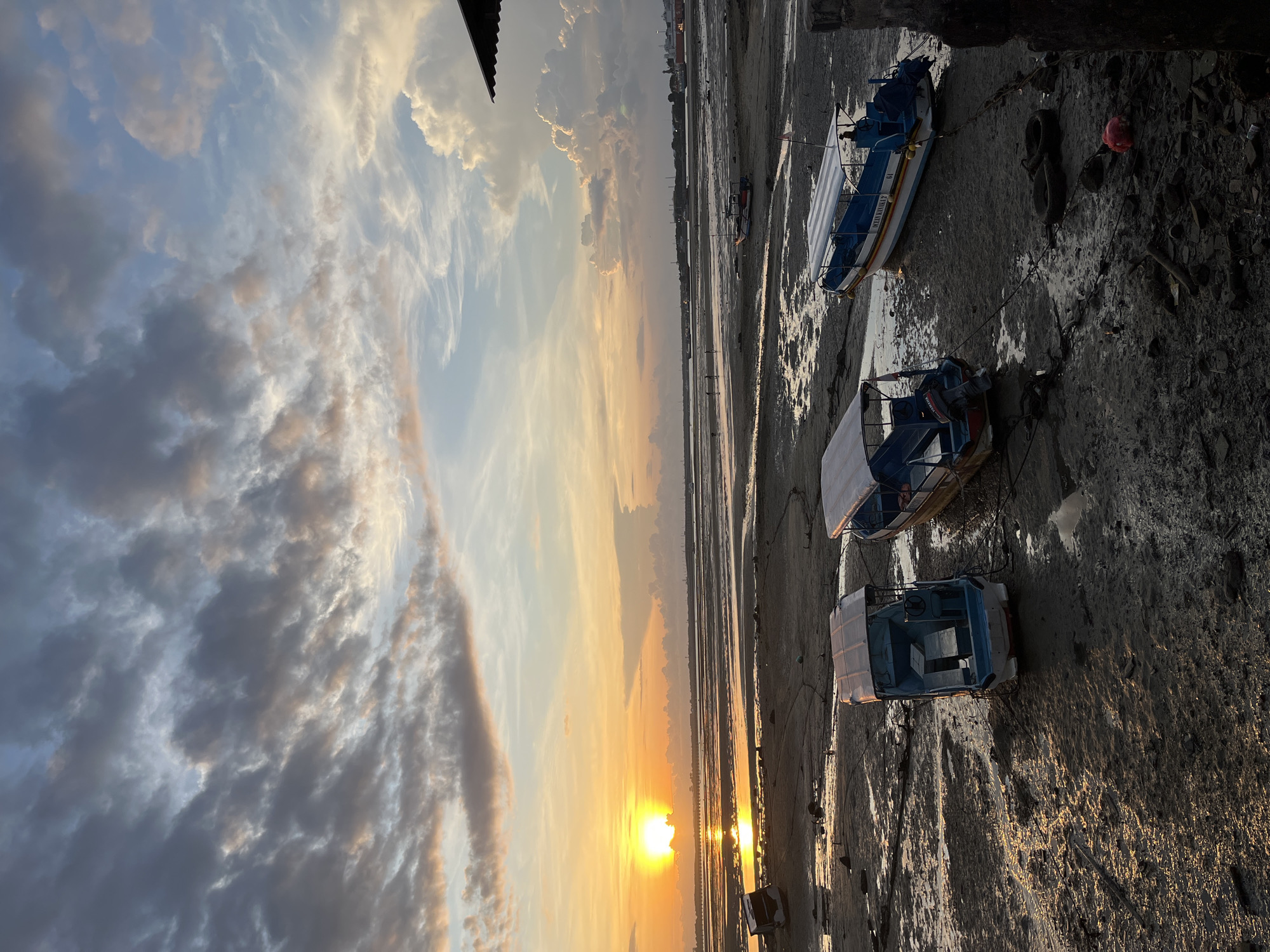 This photograph depicts a tranquil sunset over a muddy tidal flat, likely in Kuta Selatan, Indonesia. The sun, a brilliant golden orb, is low on the horizon, casting warm, radiant light across the scene. Its reflection creates a shimmering path on the wet, exposed mud and any lingering pools of water.
The sky is filled with an array of clouds. Those closer to the setting sun are illuminated with soft hues of orange, yellow, and pink, blending into the blues and grays of clouds higher up and further from the horizon.
In the foreground and mid-ground, the extensive muddy shoreline is revealed by the low tide. Several small, open boats are beached or moored on this exposed bank. On the right side of the frame, a blue and white boat clearly displays the text "OCEAN FAMILY 99" along its side, with the number "19" visible on its bow. Another small boat, closer to the foreground, features an outboard motor with the "YAMAHA" brand logo. Scattered on the muddy ground near the boats are items such as old tires and a distinctive red object.
No people are visible in the image, contributing to the quiet and serene atmosphere of the evening. The scene captures the calm beauty of dusk in a coastal or estuarial environment.