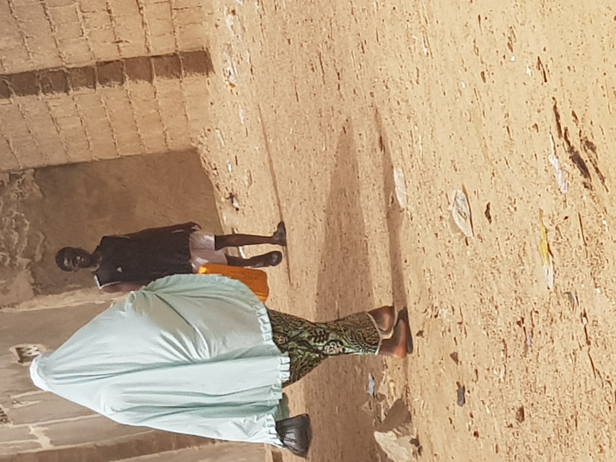The image depicts two individuals in an outdoor setting. The foreground shows a woman dressed in traditional attire: a light blue robe that covers much of her body, patterned green pants, and black shoes. Behind her is a man wearing a dark T-shirt and light-colored shorts. They are standing in a sandy, arid area. The background includes a building made of light-colored bricks. The scene's overall appearance suggests a location within the city of Dutsinma B, Nigeria.