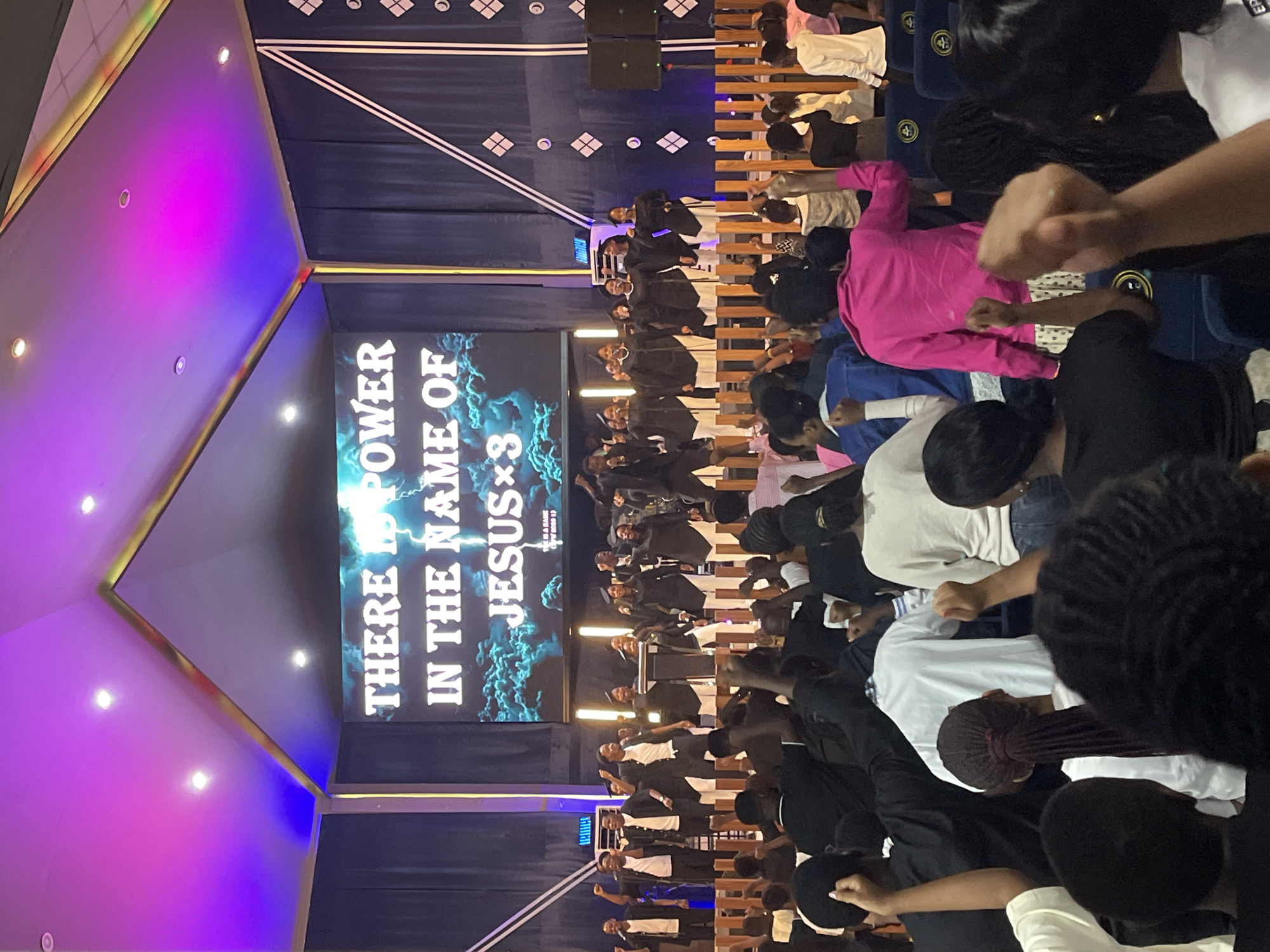 The image shows a large group of people inside a building, likely a church, in Dar es Salaam, Tanzania. They are standing in rows facing a stage with a large screen displaying the text "THERE IS POWER IN THE NAME OF JESUS ×3". Some individuals are raising their fists in the air. The people on stage appear to be a choir dressed in black robes with white shirts. The interior design features geometric shapes and purple and blue lighting.
