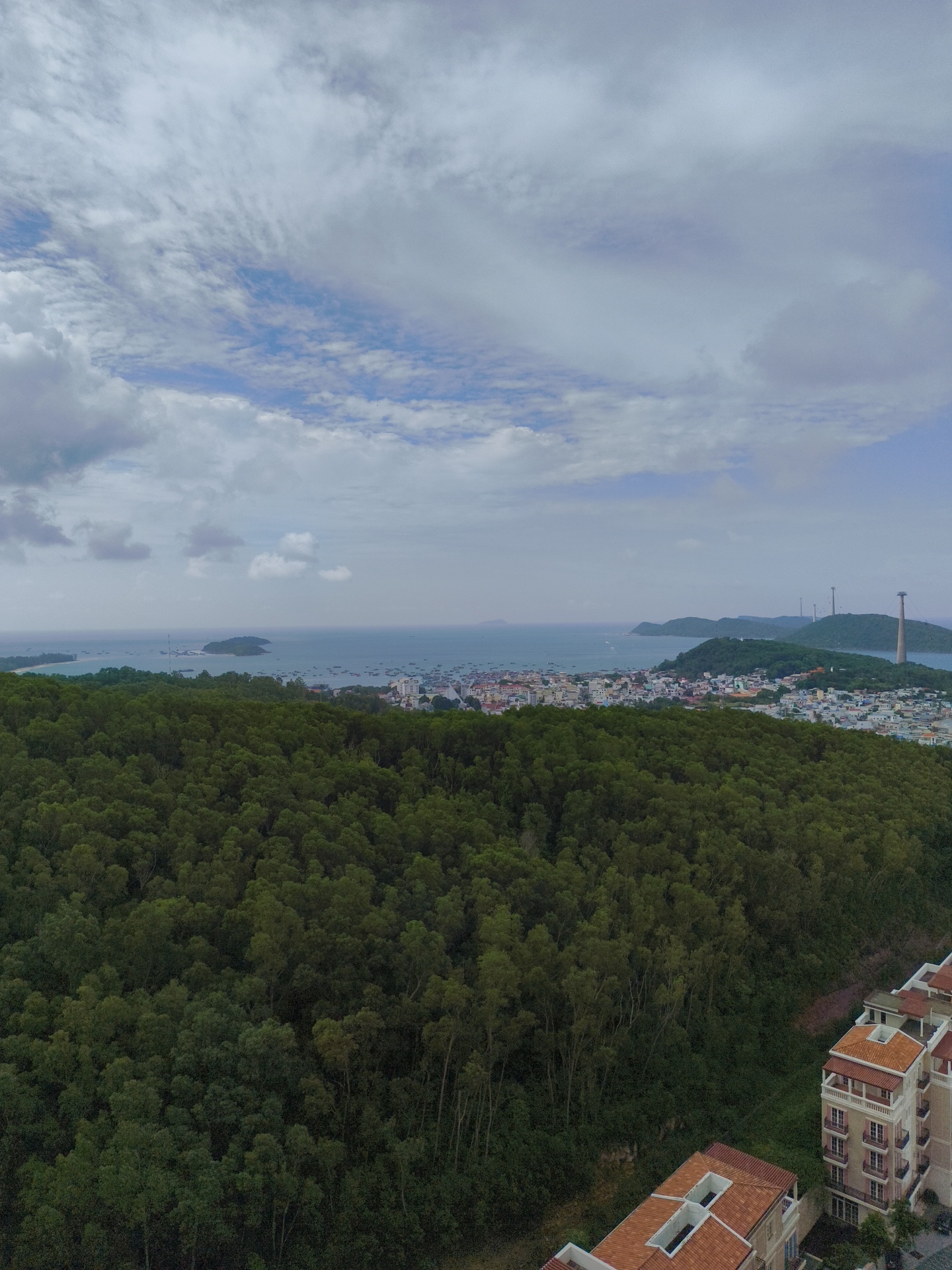 The image is an aerial view overlooking a densely forested hillside on the island of Phú Quốc, Vietnam. In the foreground, two modern apartment buildings with terracotta-tiled roofs are visible. The hillside is covered in a thick canopy of green trees. Beyond the trees, a coastal town with numerous buildings stretches along the shoreline. The town is situated on a bay filled with many small boats. In the distance, several islands and peninsulas are visible, and a tall tower with a circular structure at its top stands on one of the hills. The sky above is partly cloudy, with patches of blue visible through the white and gray clouds. The overall scene depicts a tropical landscape where nature and urban development meet the sea.