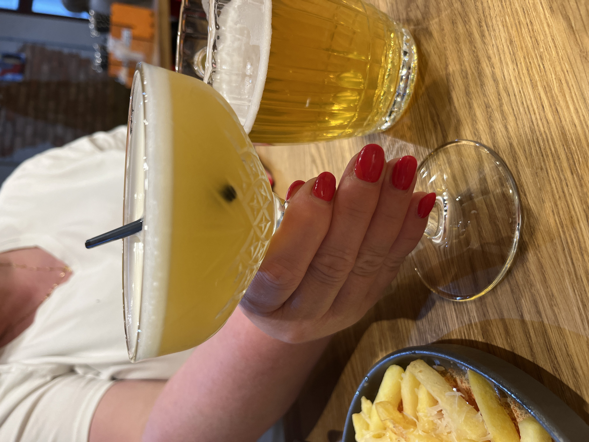 A person with red nail polish is holding a cocktail glass filled with a yellow beverage, topped with foam and a single black seed. To the right of the cocktail is a glass of beer. In the foreground, a dish of french fries with grated cheese is partially visible. The scene appears to be indoors, likely at a restaurant or bar in the city of Novo-Nikolskoye, Russia, given the context of the query.
