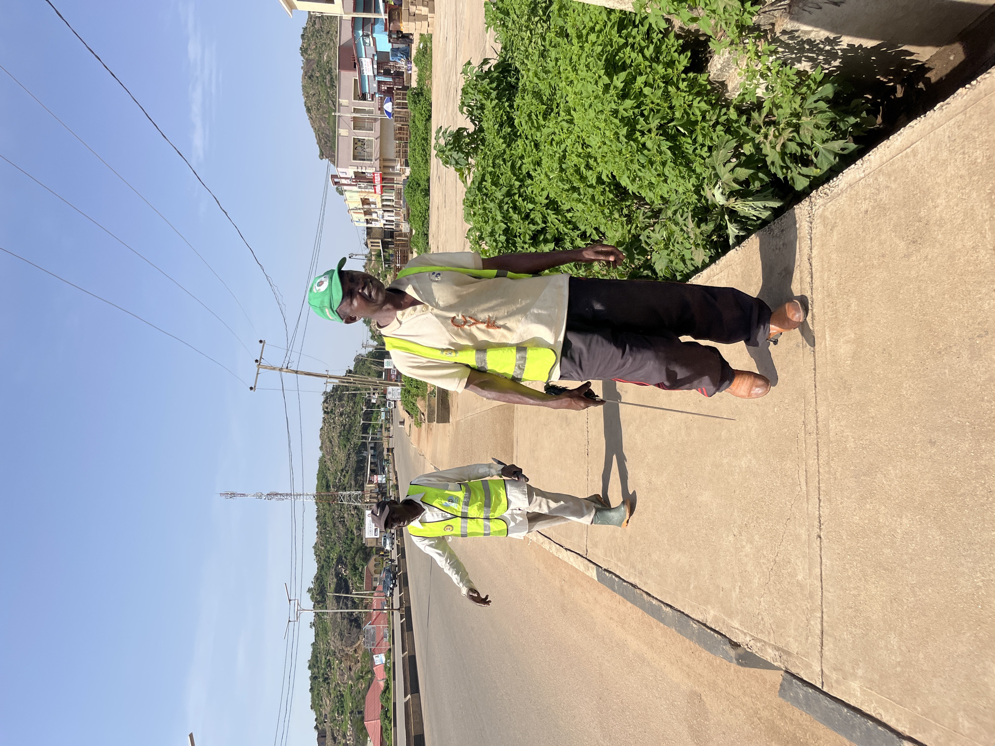 Two individuals, dressed in reflective vests and casual attire, walk along a concrete sidewalk in Gura Top, Nigeria. The individual on the right wears a green cap and a light-colored shirt with a reflective vest, while the person on the left wears a white shirt, white pants, green rubber boots, and a reflective vest. Both appear to be walking in the same direction. The environment consists of a road to the left, a sidewalk where the individuals are walking, and dense green vegetation to the right. In the background, buildings and a transmission tower are visible, with a hilly landscape and a clear sky. The lighting suggests it is daytime.