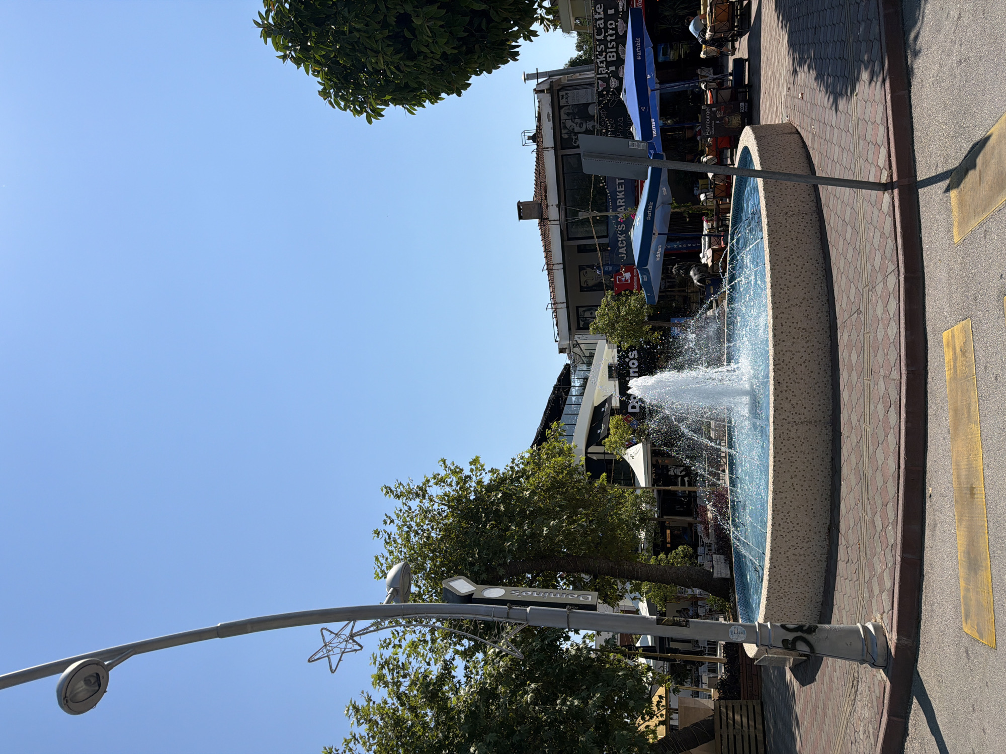 In the city of Kemer, Turkey, a circular fountain with bright blue water is the central feature of this outdoor scene. The fountain is surrounded by a decorative stone border and is situated on a paved area with a reddish-brown and grey patterned surface. A lamppost with a decorative star-shaped element stands to the left of the fountain. Beyond the fountain, several outdoor cafes and shops are visible, with patrons seated at tables under blue umbrellas. Signs for businesses such as "Jack's Market," "Domino's," and "Jack's Cafe Bistro" are displayed. Trees provide shade in the background, and the clear blue sky suggests it is a sunny day. A yellow pedestrian crossing is visible in the foreground.