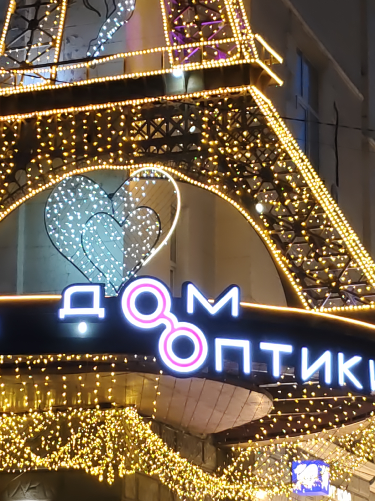 An exterior shot captures a brightly lit building facade decorated with an Eiffel Tower-inspired illuminated structure at night. The Eiffel Tower replica is adorned with numerous golden lights, creating a dazzling effect. In the center, two stylized white hearts made of lights are displayed. Below the hearts, a neon sign reads "ДОМ ОПТИКИ" in Russian, which translates to "House of Optics." The foreground is filled with a cascade of small, twinkling golden lights, suggesting a festive or commercial setting. The overall scene conveys a sense of celebration or a well-decorated shopfront. The location is the city of Rostov-on-Don, Russia.