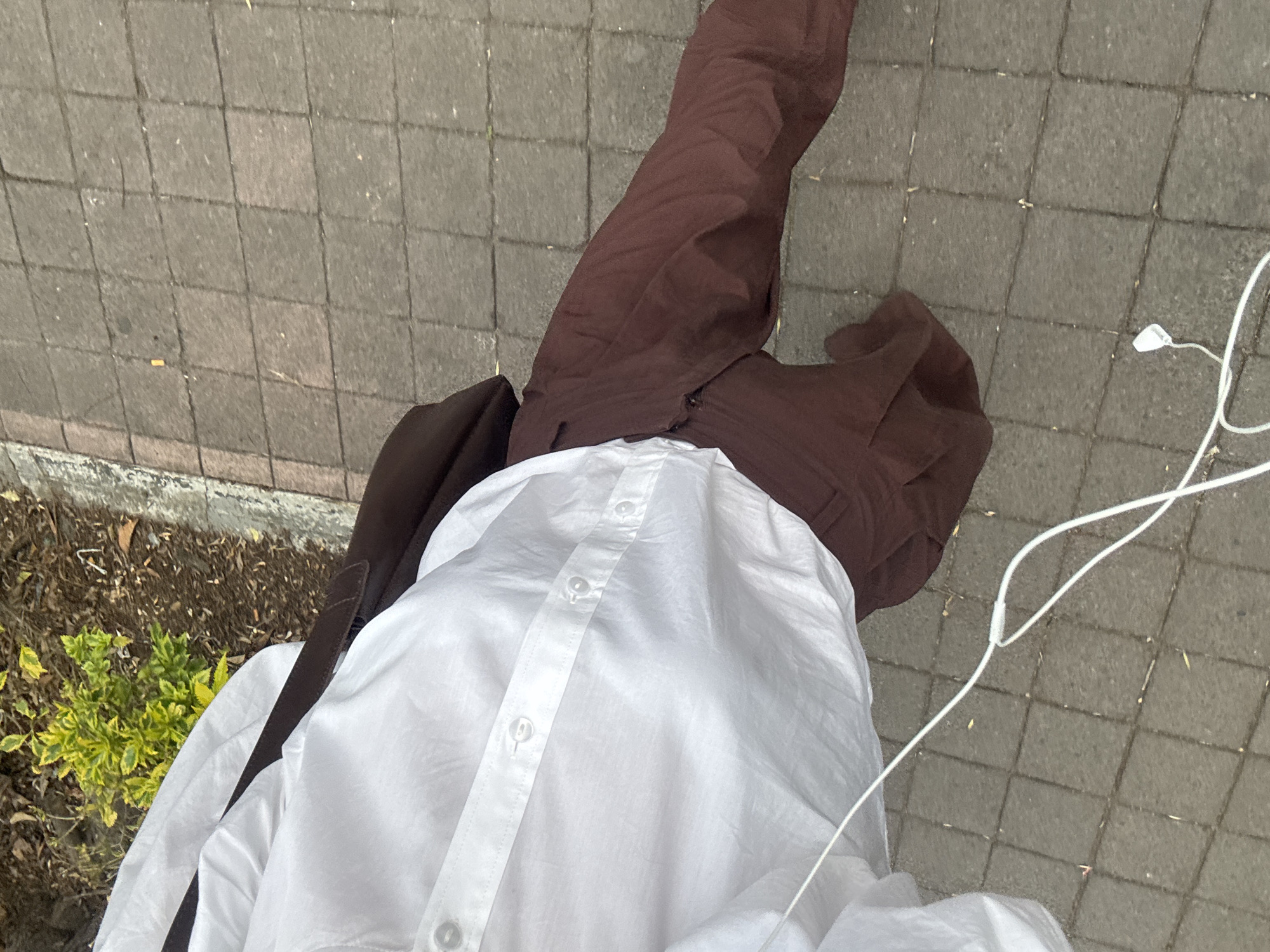 The image presents an overhead view of a person standing outdoors, likely on a sidewalk or paved area. The individual is wearing brown pants and a white button-down shirt. A brown jacket is draped over their shoulder. White earbud cables are visible, suggesting the person is listening to music or a podcast. The ground is paved with gray rectangular stones. A small patch of greenery is visible along the edge of the pavement.