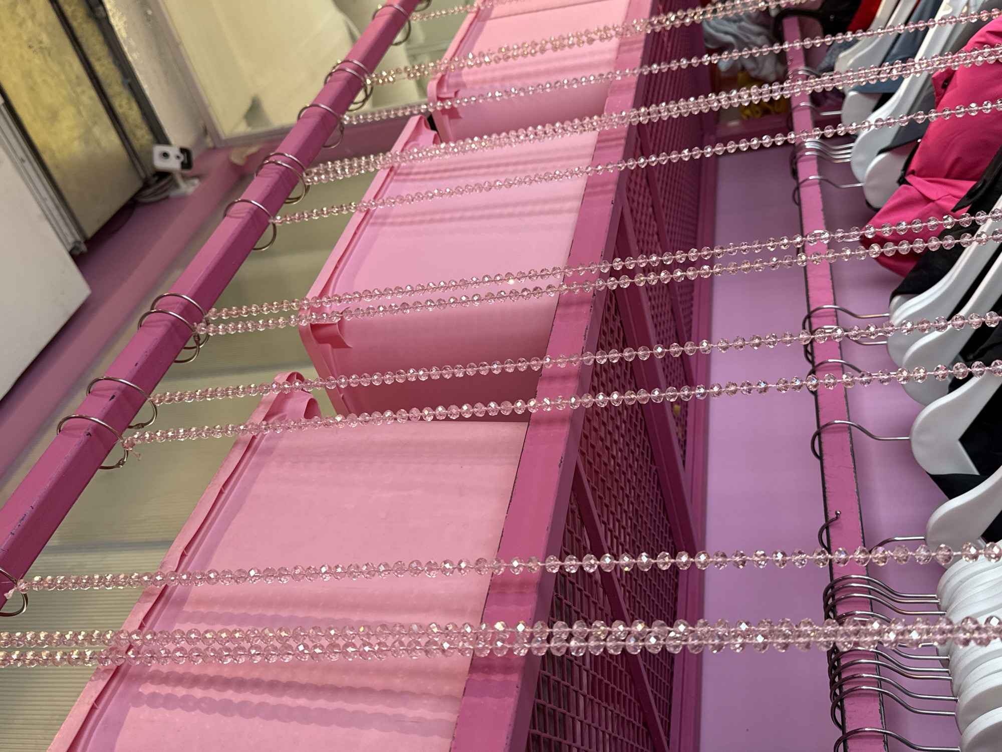 The image shows a close-up of a pink clothing rack. Three pink rectangular containers are stacked vertically on the left side of the rack. Strands of pink beads hang horizontally between the containers and extend to the right side of the rack, where clothing hangs on white hangers. The background is a solid pink wall. The location is not discernible from the visual information provided.