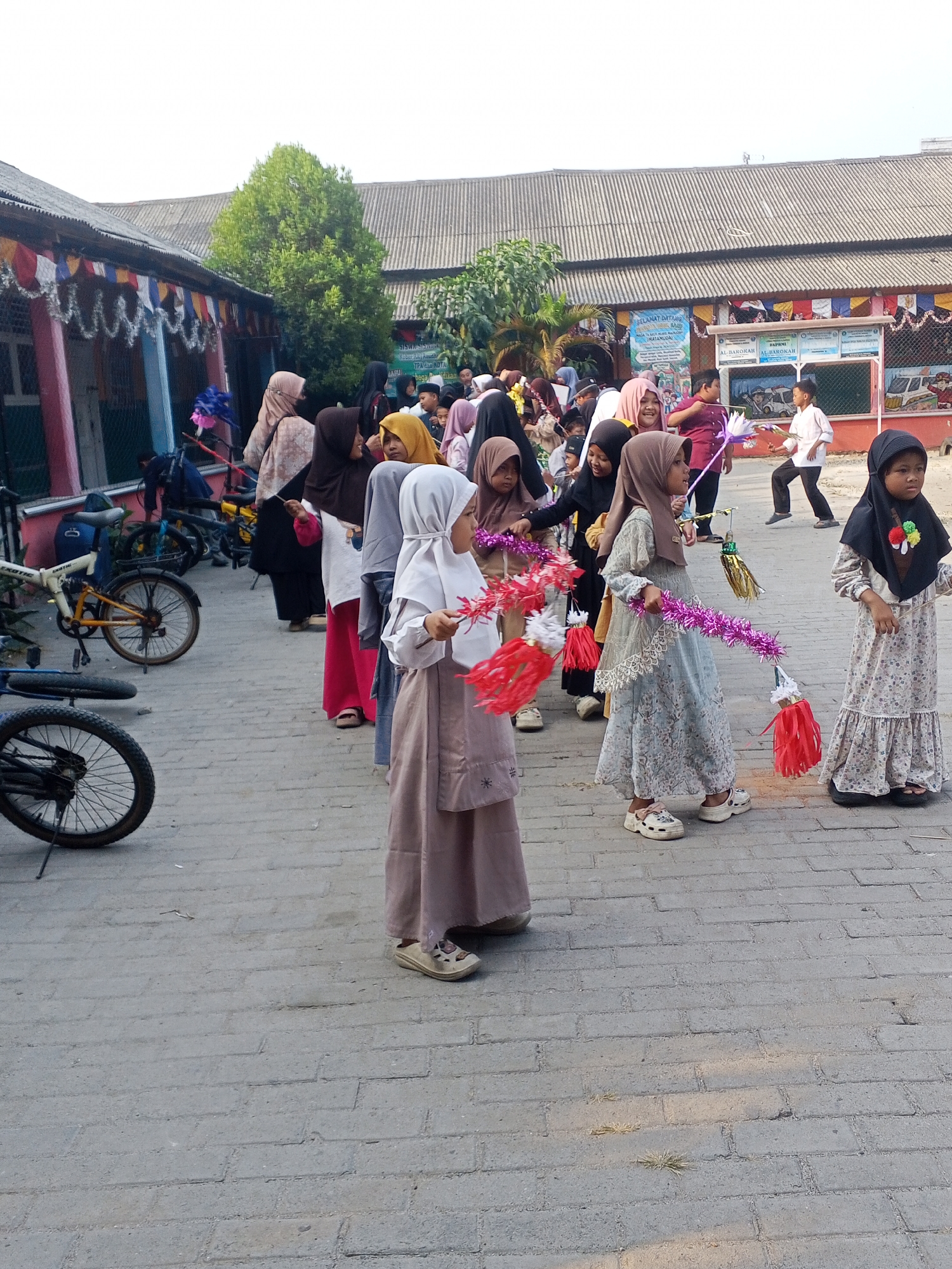 In Ciruas, Indonesia, a group of young girls wearing hijabs are gathered outdoors, likely in a schoolyard, as indicated by the building in the background decorated with colorful banners and what appear to be school-related posters. The girls are holding festive decorations, some resembling sparklers or streamers, in shades of pink, red, and gold. They are dressed in traditional Islamic clothing, with some wearing long dresses and hijabs in various colors like beige, brown, and light blue. The ground is paved with interlocking gray bricks. Several bicycles are parked in the background, suggesting a common mode of transport in the area. The atmosphere appears to be one of celebration or a school event, with the children looking engaged and possibly anticipating an activity. The lighting suggests it is daytime, with natural sunlight illuminating the scene.