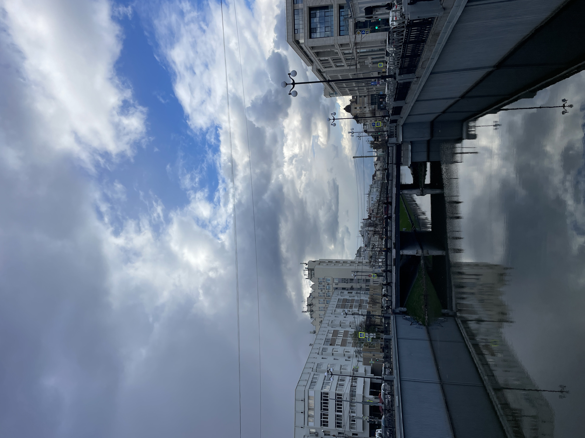 The image captures an urban waterside scene, likely a canal or river, lined with buildings and crossed by a bridge, under a partly cloudy sky. The perspective is from one side of the waterway, looking across to the opposite bank and down the length of the channel.
In the upper portion, the sky dominates, featuring a mix of bright white and dark grey clouds alongside patches of vibrant blue. Thin utility lines are visible stretching across the sky.
Below the sky, the cityscape unfolds. Multi-story buildings of varying architectural styles, from more traditional to modern, line both embankments. One side features a taller, light-colored building with numerous windows, while the opposite side shows a mix of structures.
A grey, seemingly concrete or metal bridge spans the waterway in the middle ground. Embankments flank the water, equipped with streetlights featuring distinctive spherical lamps. Cars are parked or moving along the roads on both sides of the water. Traffic signs, including yellow square signs depicting pedestrian crossings, are visible along the embankments. A small patch of green grass can be seen on one side of the water, near the bridge.
The calm surface of the water acts as a mirror, reflecting the clouds, buildings, and streetlights from above, creating a distorted yet clear parallel image.
No people are visible in the scene. The time of day appears to be daytime, though the presence of varied cloud cover suggests it could be an overcast day, morning, or late afternoon. The overall weather appears calm. The traffic signs with pedestrian crossing symbols are clearly visible. The architecture and street signage are consistent with an urban setting in Russia, aligning with the given context of Kazan. The scene presents a tranquil, everyday urban landscape.
