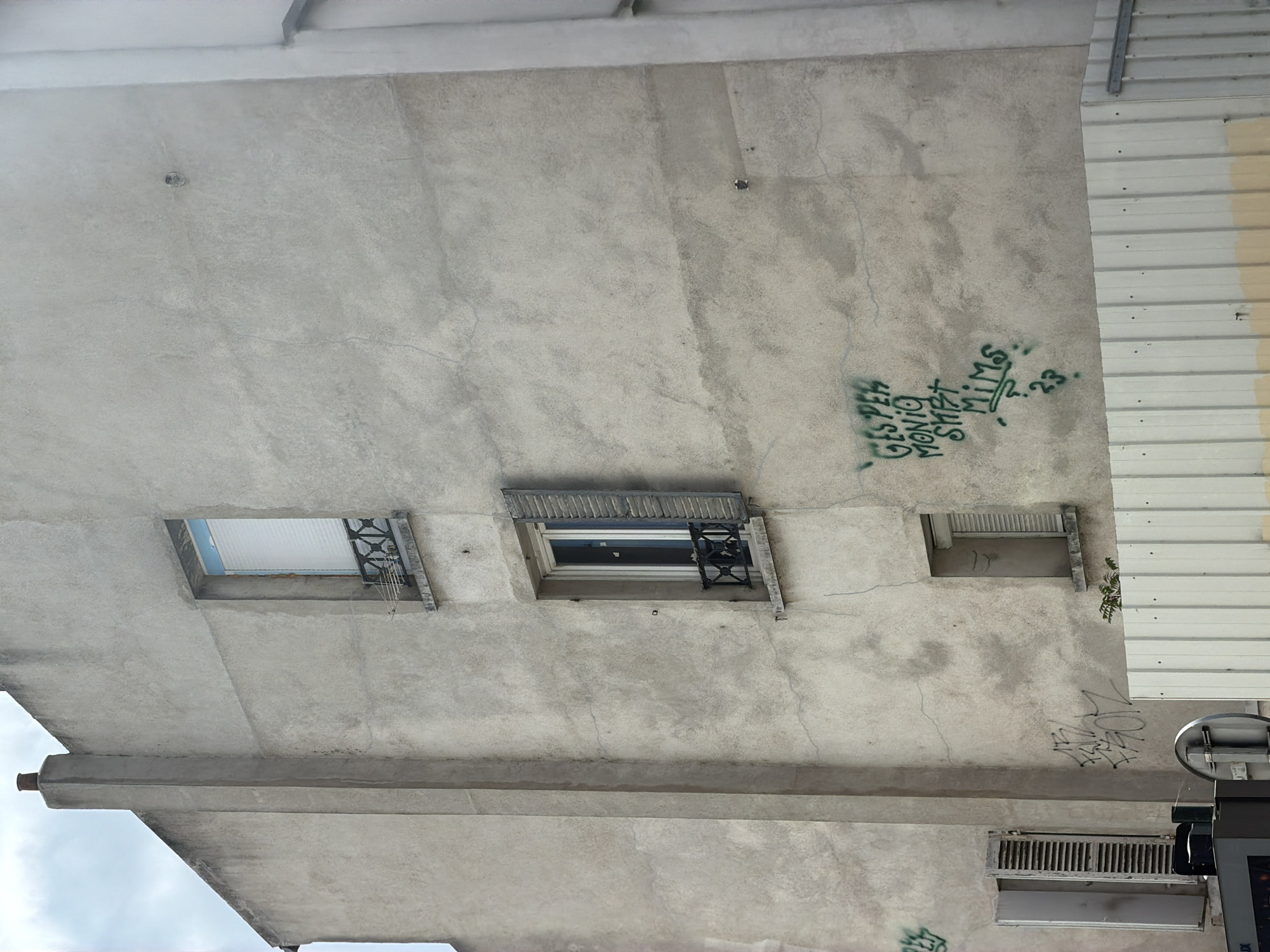 The image shows the exterior of a concrete building, viewed from below. The concrete is rough and shows signs of wear, with some cracks visible. There are three small windows or vents on the surface. The left window is open, revealing a white ceiling and some metal bars. The middle window is also open, with a view of a dark interior and a decorative metal grille. The right window is smaller and appears to be a vent.
Green graffiti is visible on the concrete. One piece of graffiti reads "GIS P EH MONTO SMET" and another reads "MI MS . 23.". There are other faint markings on the wall.
To the right of the concrete wall is a section of corrugated metal siding, painted in alternating bands of white and pale yellow.
The overall impression is of an urban or industrial setting, possibly a less-maintained part of a building. The sky is visible in the lower left corner, suggesting it's daytime, but the lighting is somewhat muted, and the clouds are not clearly discernible. The perspective is unusual, looking upwards at the underside of a structure.