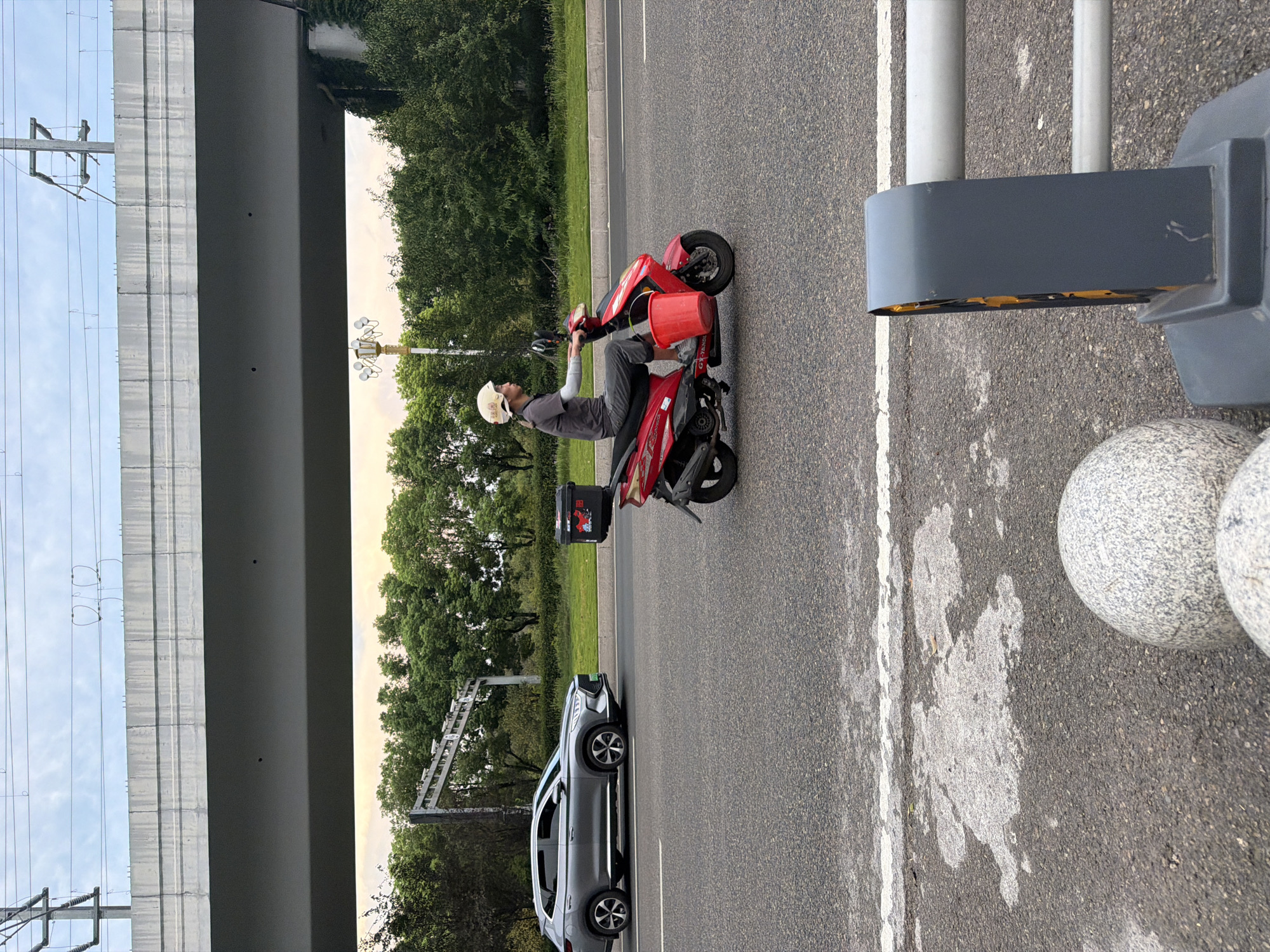 In Jiangjin, China, a person rides a red scooter down a multi-lane road. They are wearing a dark t-shirt, grey pants, a light-colored helmet, and arm sleeves. A red bucket is attached to the front of the scooter, and a black storage box is on the rear. A gray SUV is driving in the adjacent lane. Above the road, a concrete overpass is visible, with electrical wires running across the top. The background features a line of green trees and a cloudy sky. In the foreground, a guardrail and two large, speckled stone spheres are present.