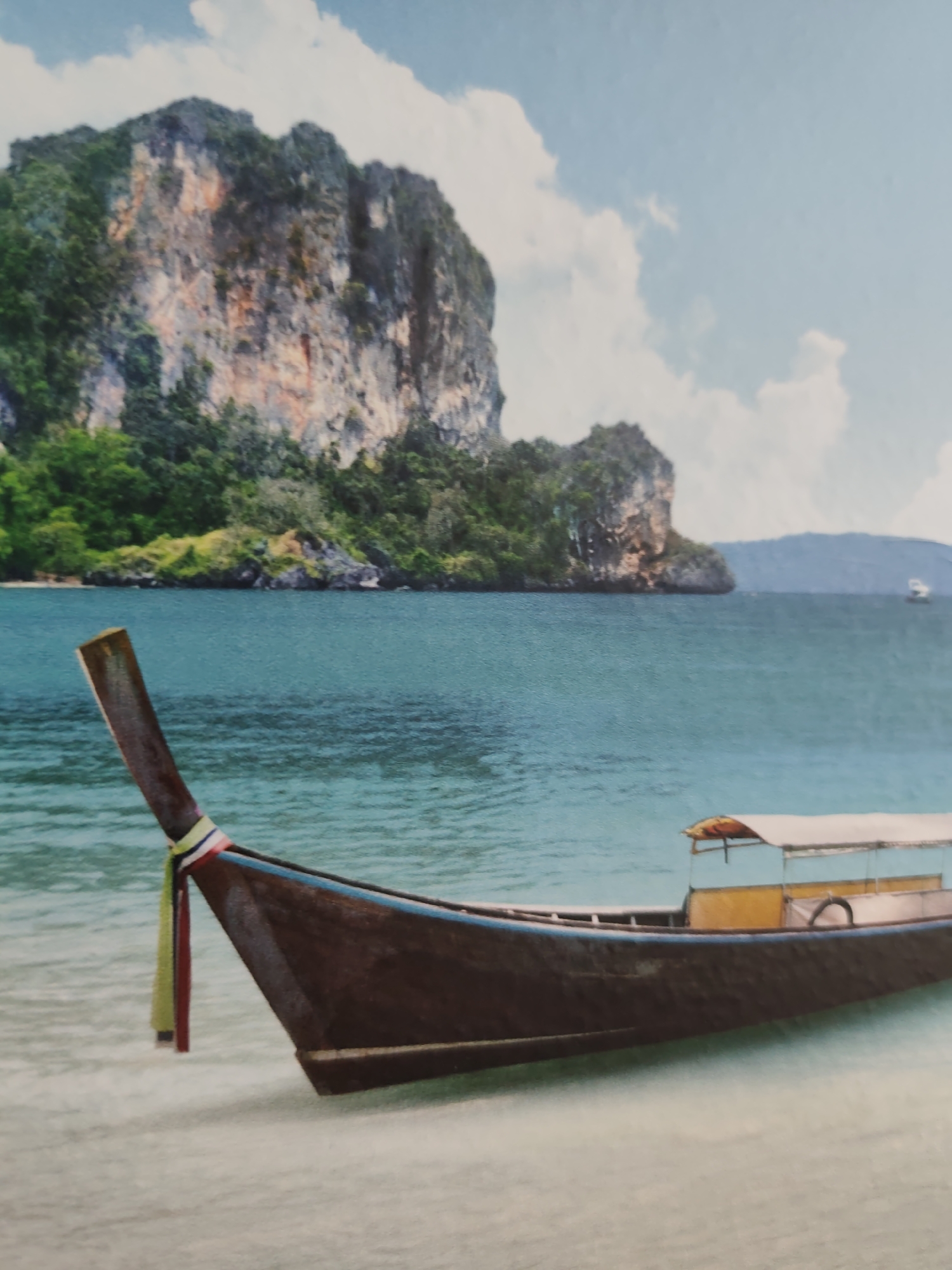 The image displays a tropical coastal landscape under a bright, partly cloudy sky. In the foreground, a dark brown wooden longtail boat is partially beached on a light sandy shore, with its hull resting in clear, shallow turquoise water. The boat's pointed prow is adorned with decorative fabric strips in red, white, and yellow. A light-colored canopy covers a section of the boat's interior, which also features yellow accents and a dark, ring-shaped object. The midground shows calm, blue-green ocean water. In the background, majestic, steep limestone karst islands, densely covered with lush green vegetation, rise prominently from the sea. A smaller, distant boat is faintly visible on the horizon to the right. No individuals are present in the scene. The depicted environment, characterized by its tropical maritime features and limestone karsts, does not correspond to the inland geography of the city of Pazardzhik, Bulgaria.