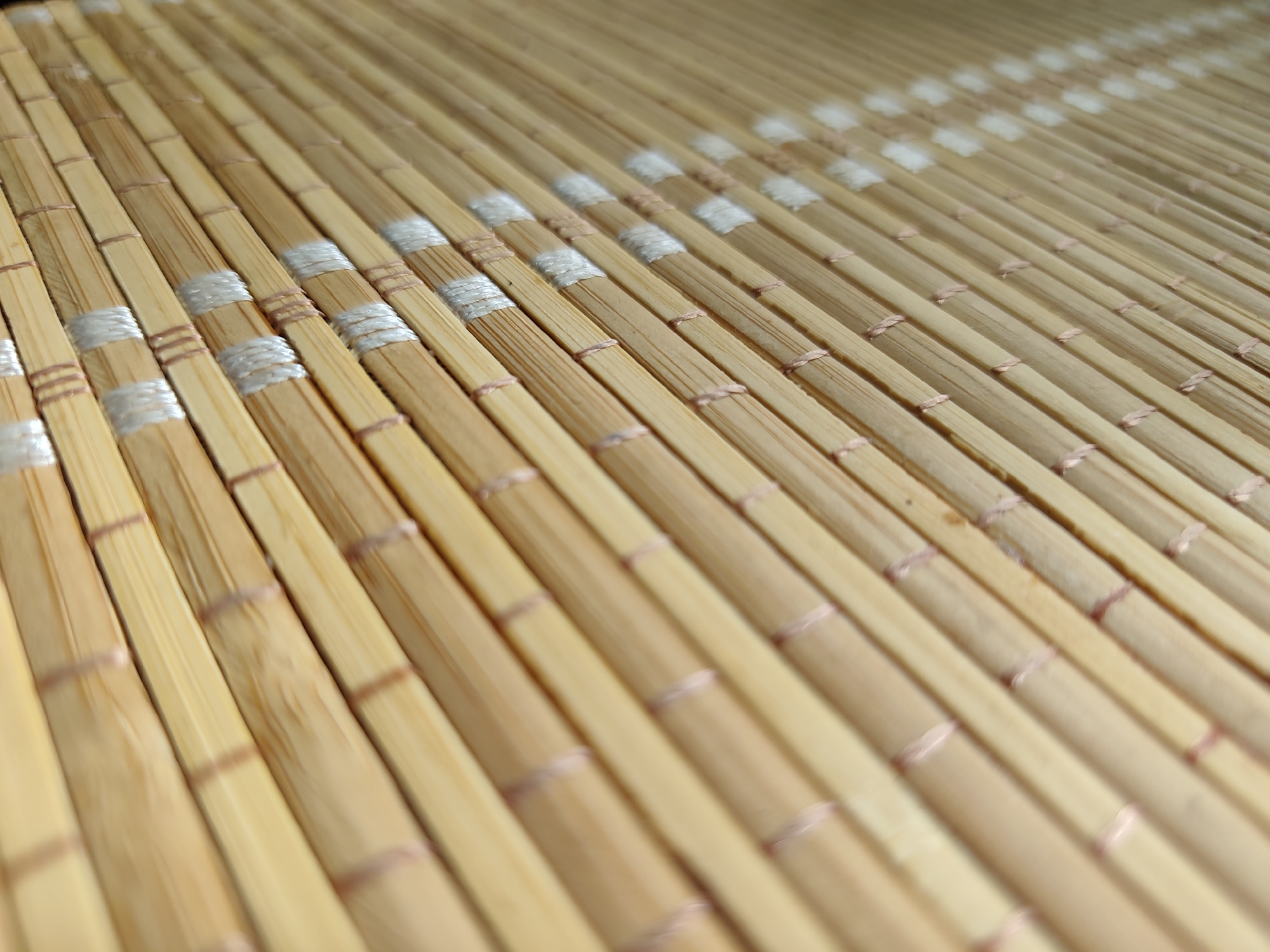 The image is a close-up, low-angle shot of a woven bamboo mat. The bamboo stalks are light brown and arranged in parallel rows. Thin, pinkish-brown twine is used to tie the stalks together at intervals. In some sections of the mat, there are also bands of white thread, possibly decorative or for reinforcement, wrapped around the bamboo stalks. The mat appears to be clean and in good condition. The perspective creates a sense of depth, with the foreground stalks in sharper focus and the background stalks progressively blurring. The lighting is natural and casts soft shadows, highlighting the texture of the bamboo and twine. There are no people or any other objects visible in the image. There is no context provided to associate this image with the city of Minsk, Belarus.
