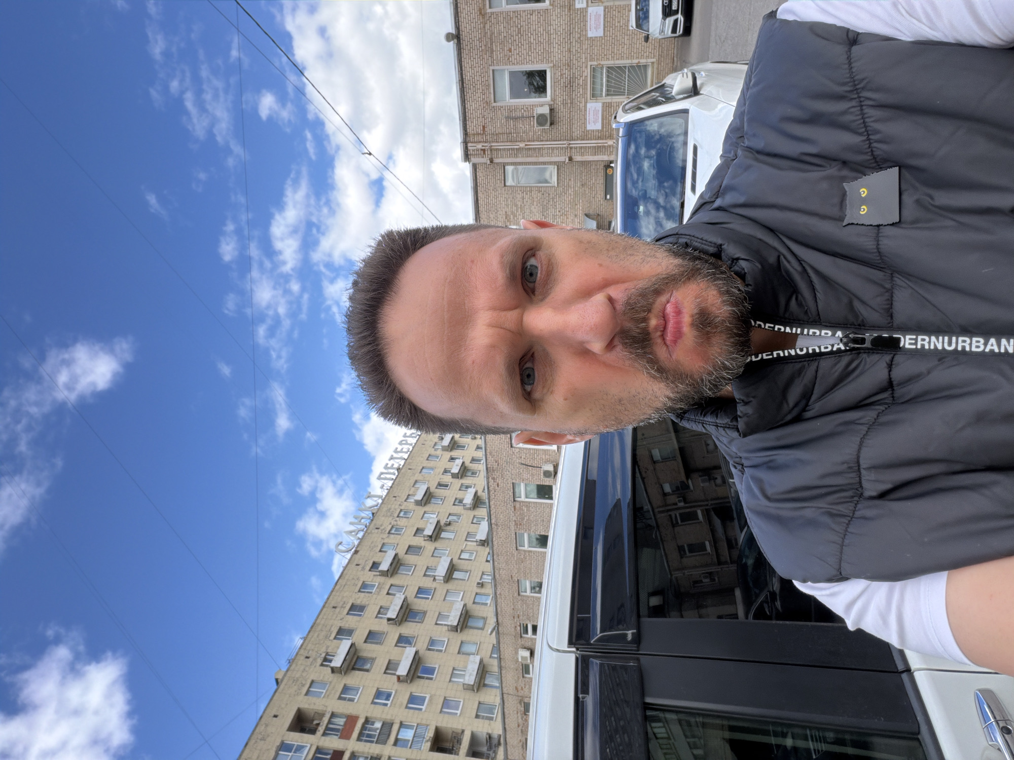 A man is taking a selfie with a building and sky visible in the background. The building has the words "САНКТ-ПЕТЕРБУРГ" on top, which translates to Saint Petersburg. The building is a multi-story residential structure with many windows. The sky is blue with scattered clouds. The man is wearing a black vest over a white shirt. A white car is partially visible in the foreground. A building with brick facade and windows is behind the white car.